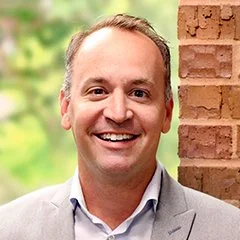 Smiling man with short hair in a light gray blazer and white shirt standing outdoors near a brick wall.