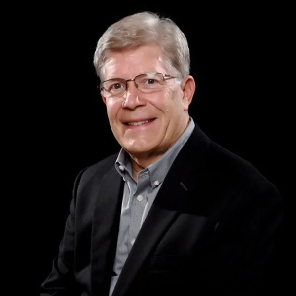 A middle-aged man with glasses and gray hair, wearing a dark suit jacket and a light gray shirt, smiling against a black background.