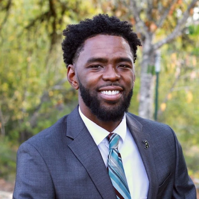 A smiling man in a gray suit with a tie, standing outdoors with trees in the background.