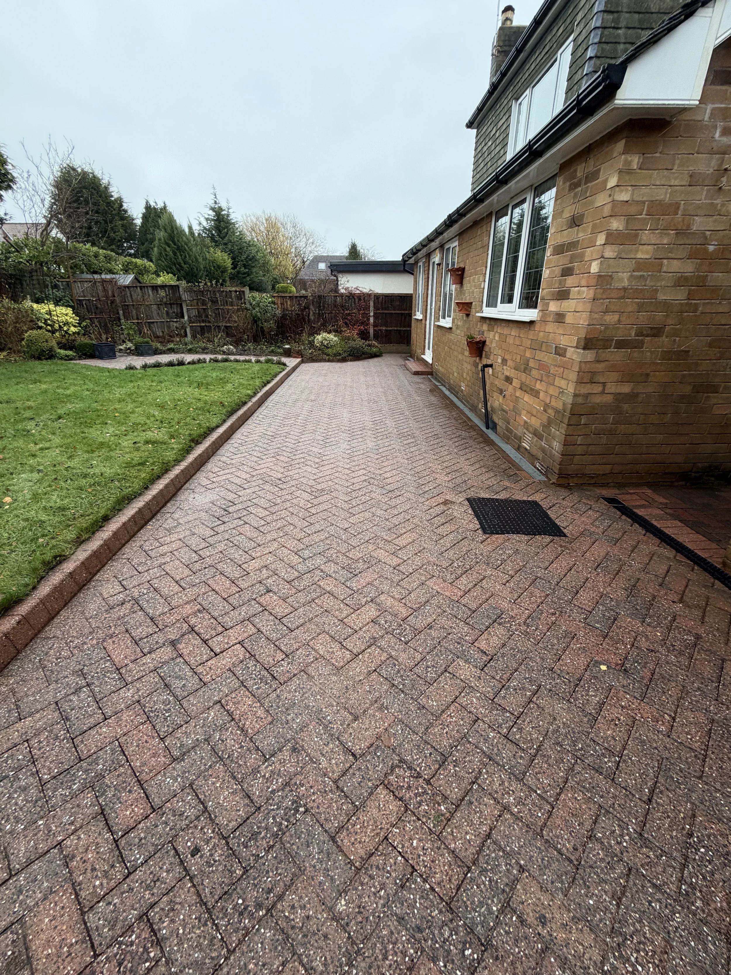 A brick patio with a herringbone pattern next to a house with a brick exterior and multiple windows. The patio is bordered by a garden with grass, bushes, and small trees, with a wooden fence in the background.