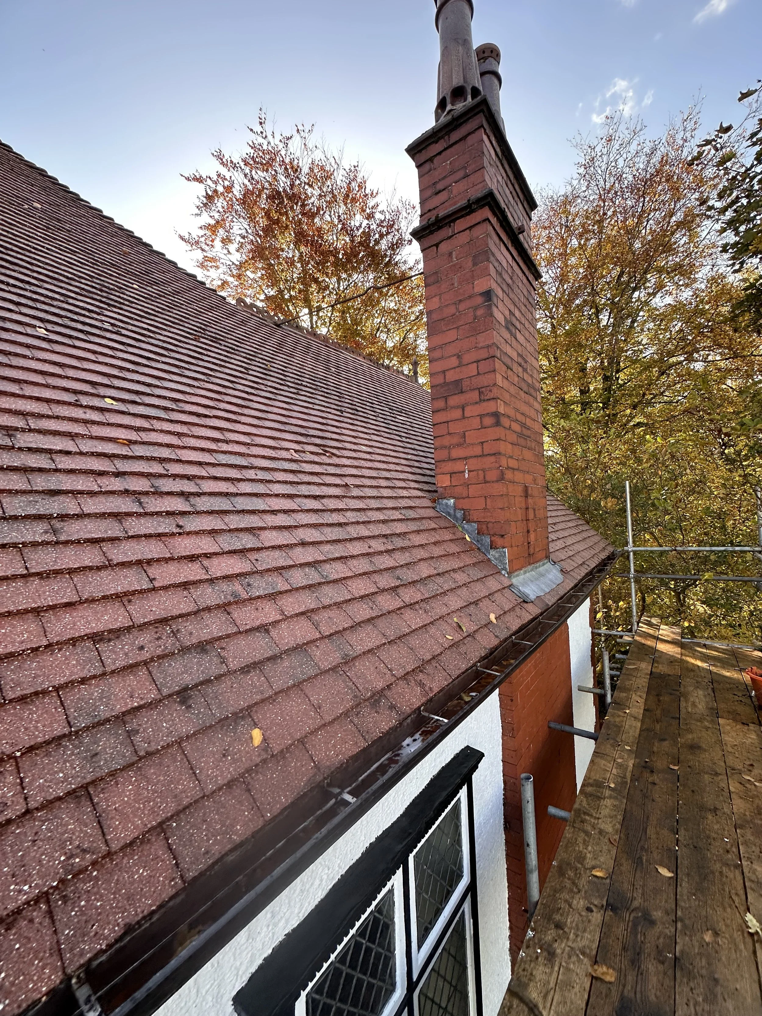 View of a red brick chimney and roof on a house with scaffolding and wooden planks, surrounded by autumn trees.