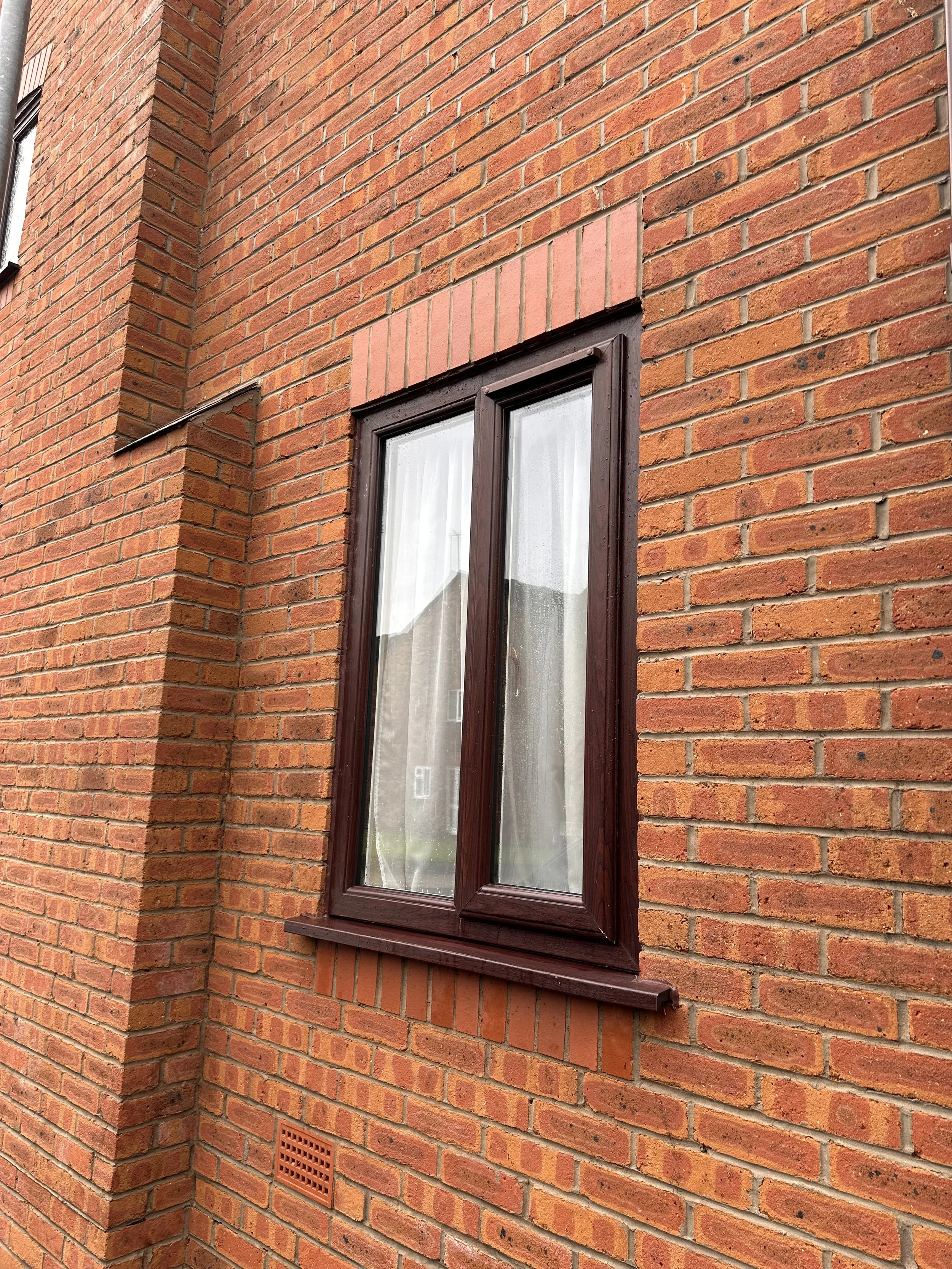 A brick house exterior with a window featuring a dark wood frame and a brick arch above it.