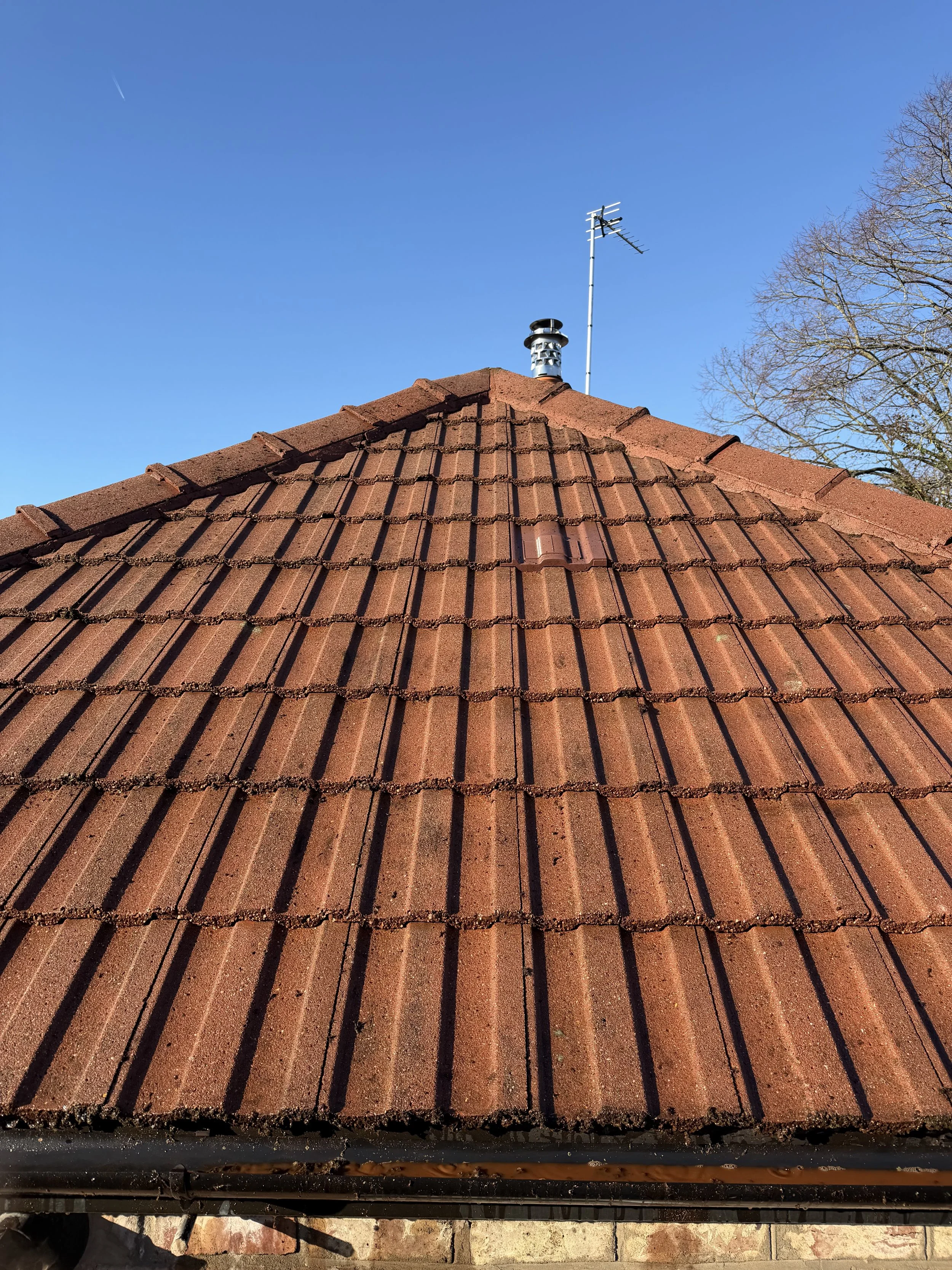 Close-up of a red tiled roof with a chimney and an antenna against a clear blue sky, with a tree in the background.