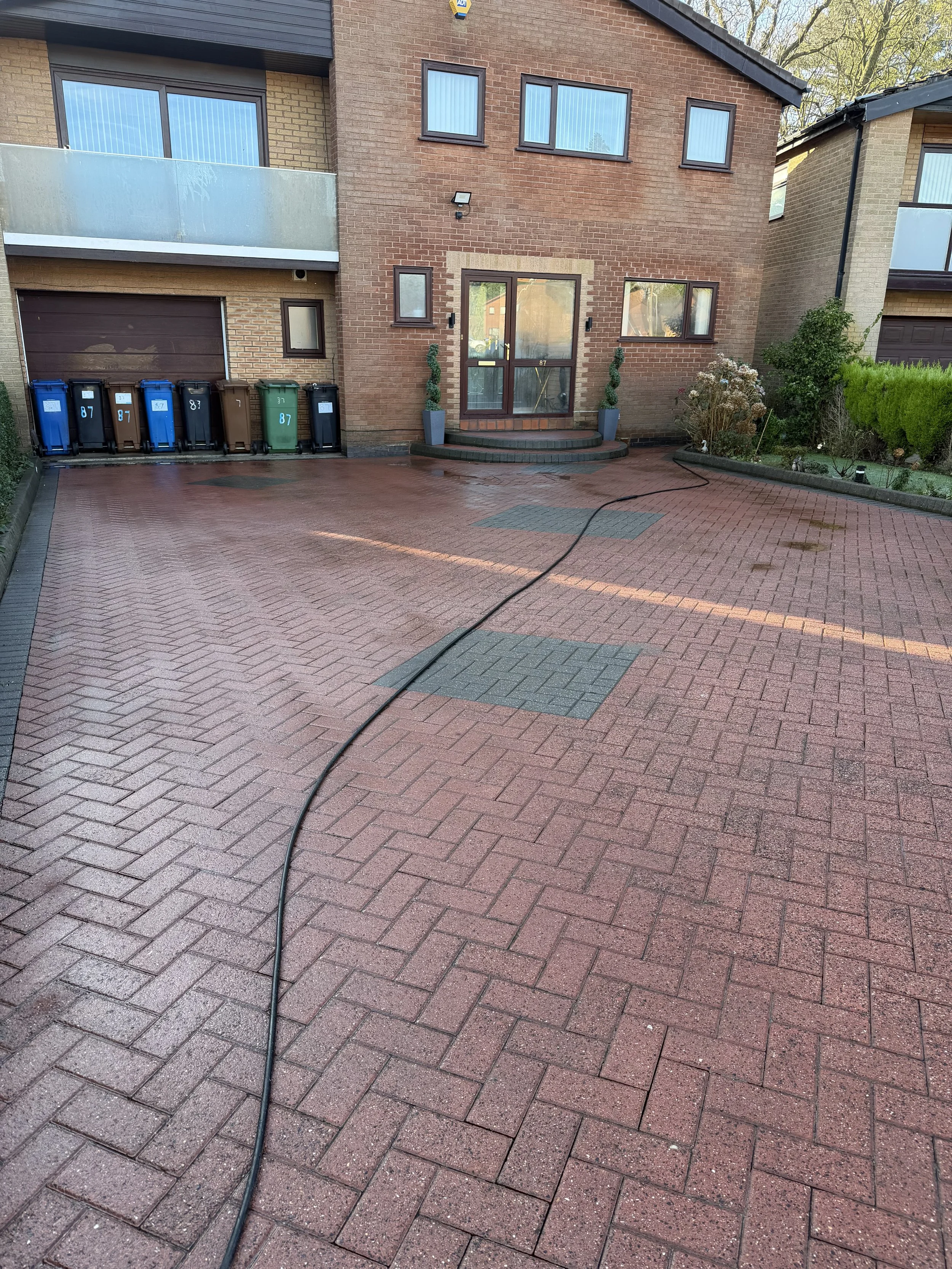 Clean, wet brick driveway in front of a modern house with a glass door, potted plants, and garbage bins to the left.