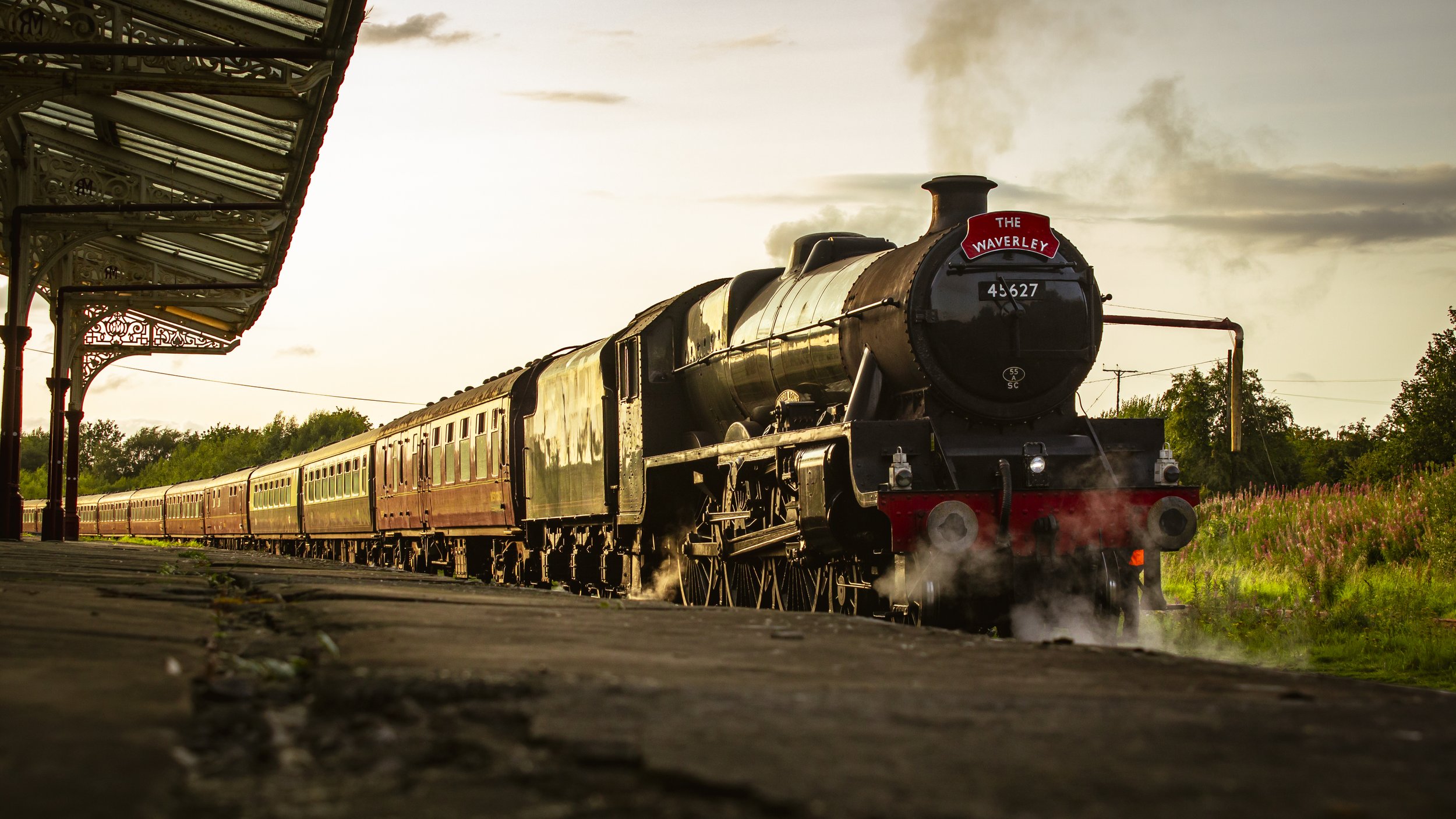 A vintage steam train pulls away from a station platform at sunset, with steam rising from its wheels and smoke from its chimney, under a sky with clouds.