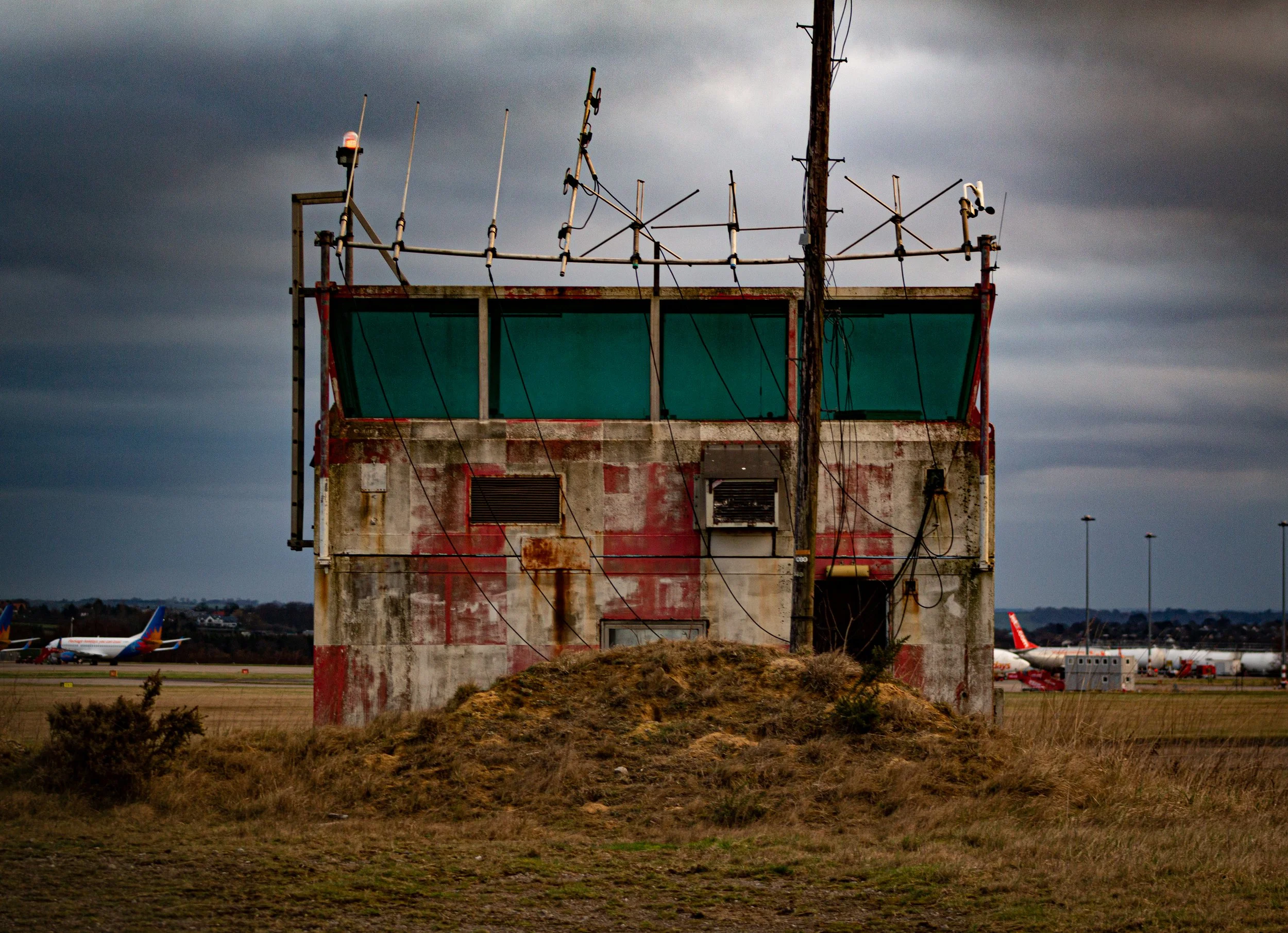 An old, weathered control tower building at an airport with rusted exterior and antennas on top. In the background, several airplanes are parked on the tarmac under a cloudy sky.