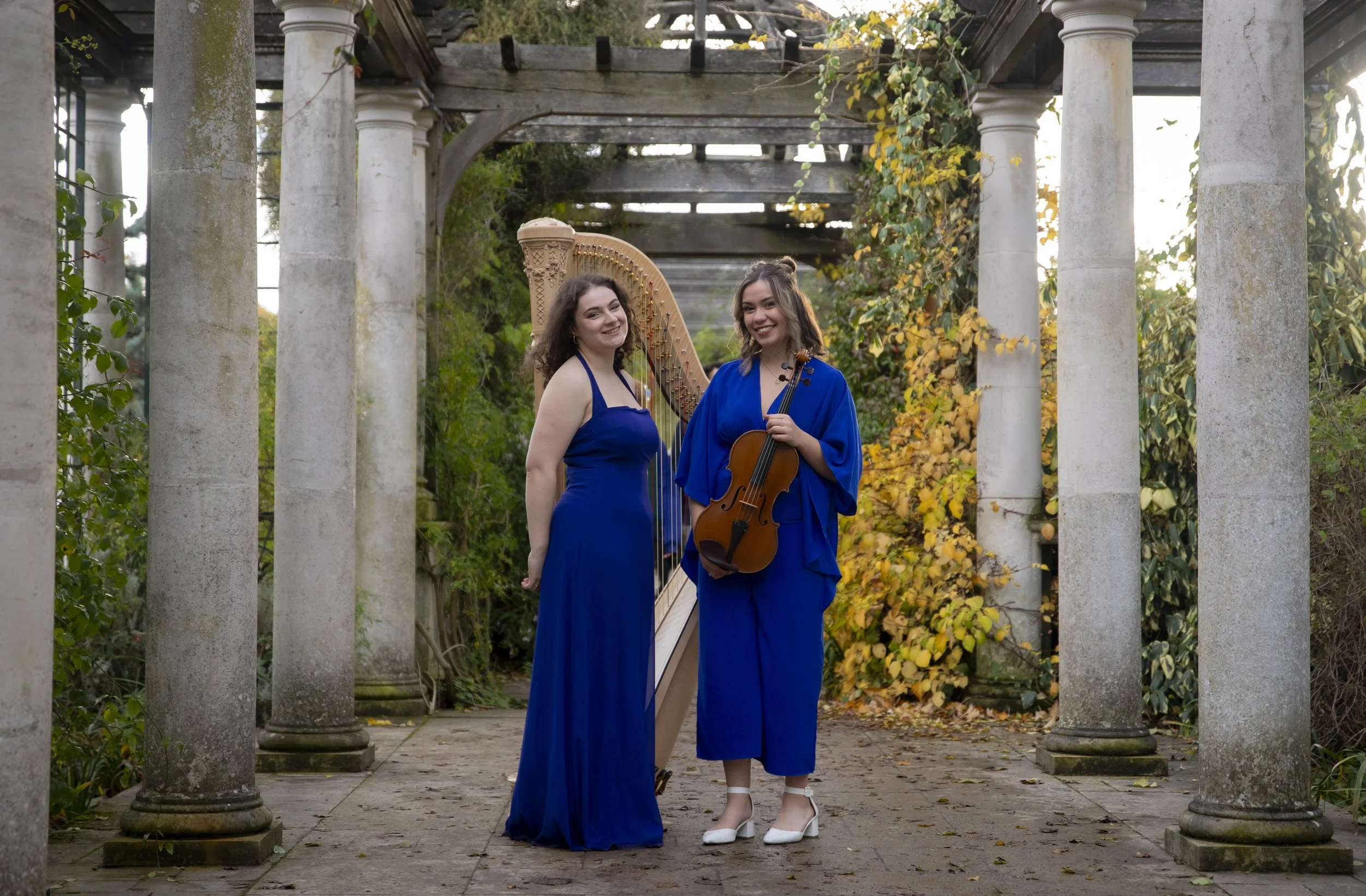 Two women in blue dresses standing in an outdoor garden with stone columns and autumn foliage. One woman is holding a violin, and the other is standing next to a harp, both smiling at the camera.