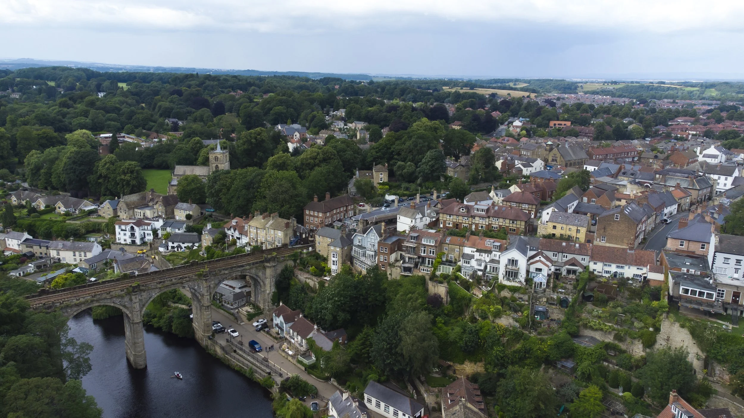 Aerial view of a small town with a river, a stone bridge, houses, a church, and lush green trees.