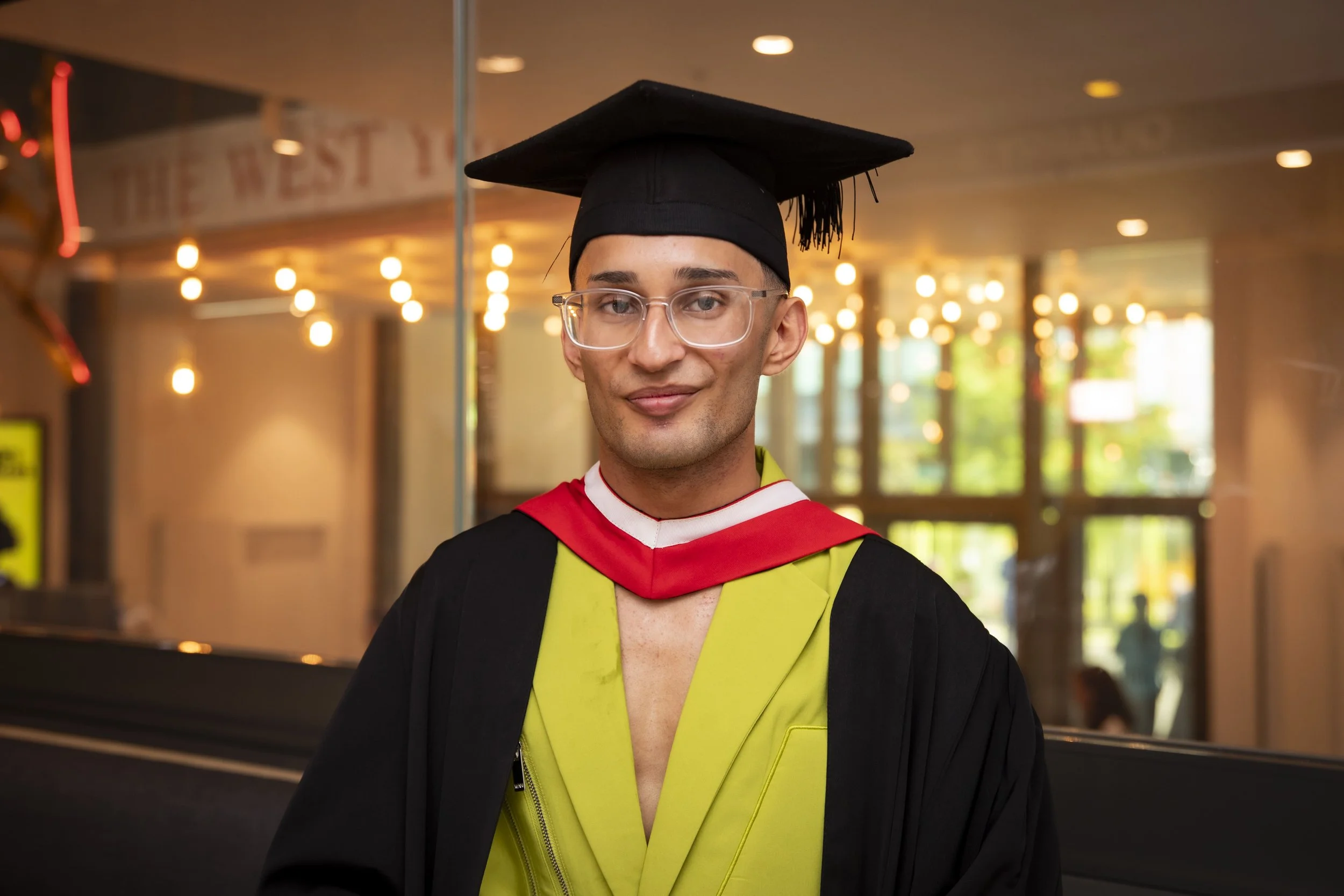 Graduate in black cap and gown with yellow and red stole, standing indoors by glass wall, smiling.