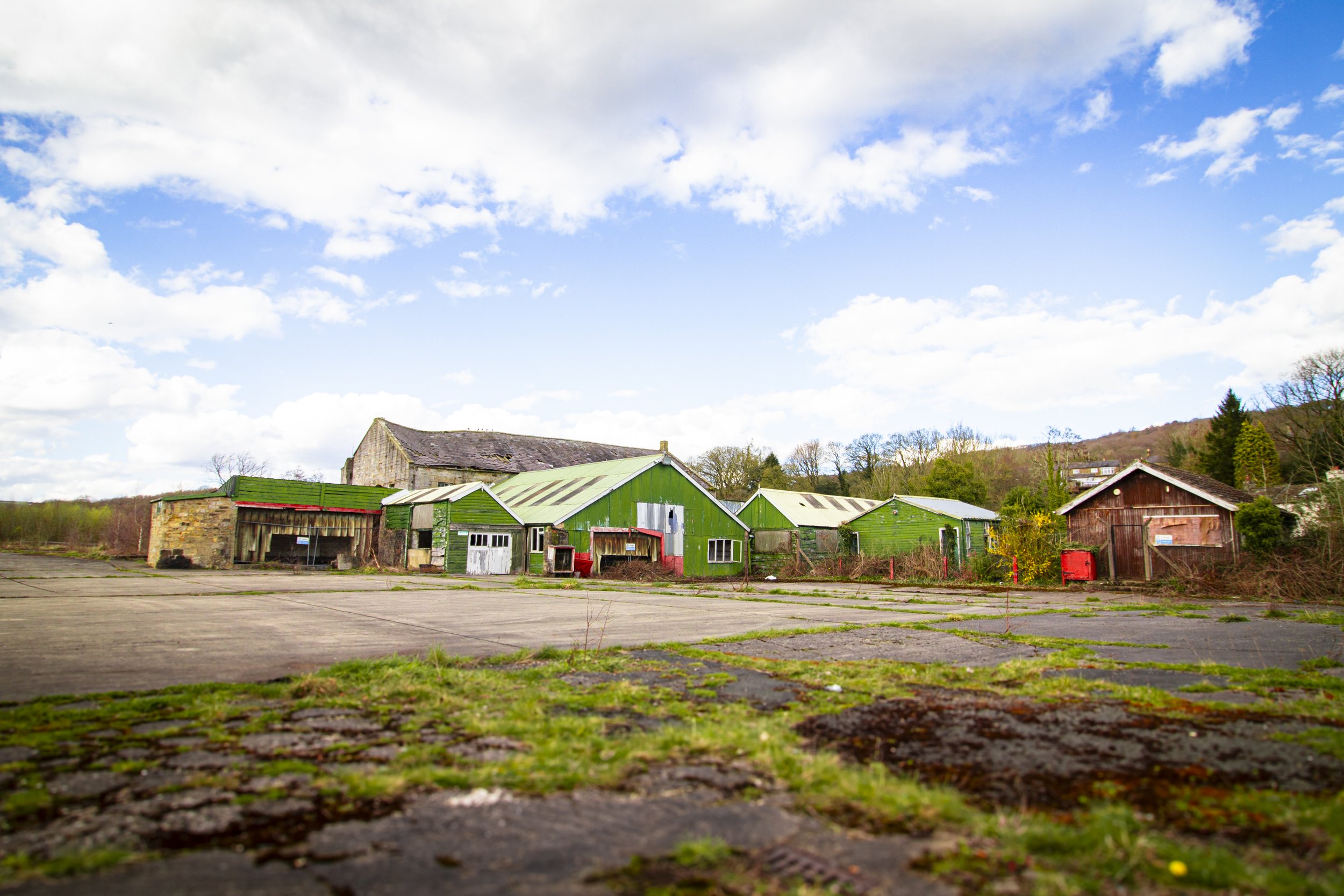 A rural scene with a grassy parking lot and a cluster of weathered green and brown buildings under a partly cloudy sky.