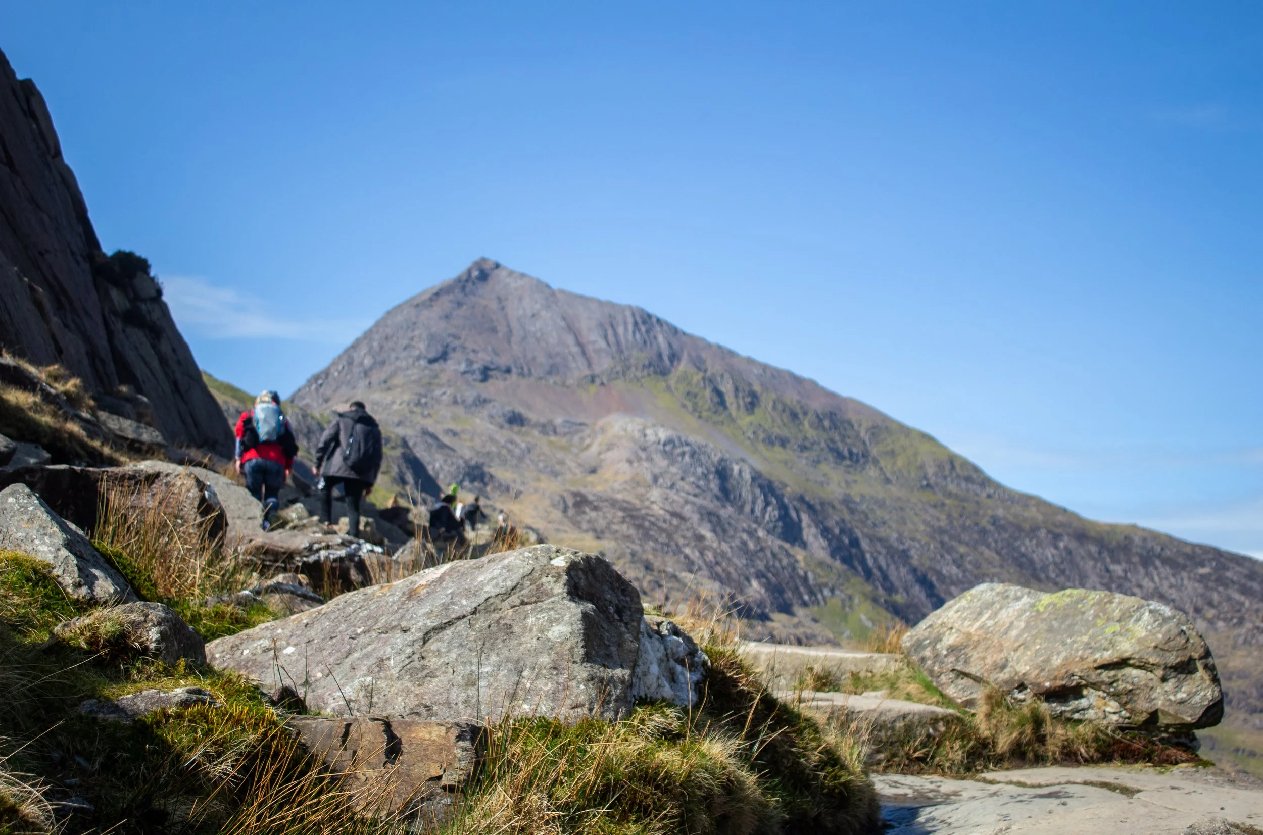 Hikers walking on a rocky trail in a mountain landscape with a clear blue sky.