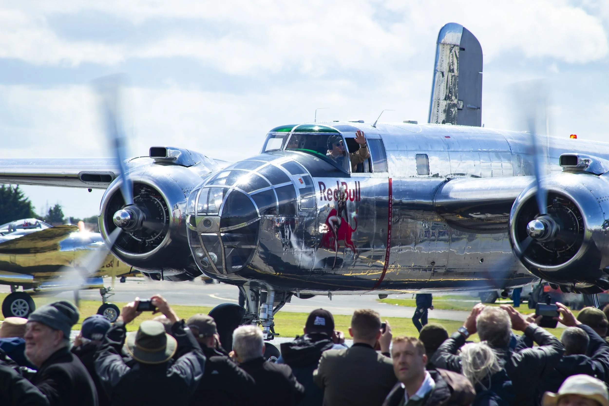 Silver aerobatic aircraft with Red Bull logo, surrounded by crowd taking photos at an airshow.