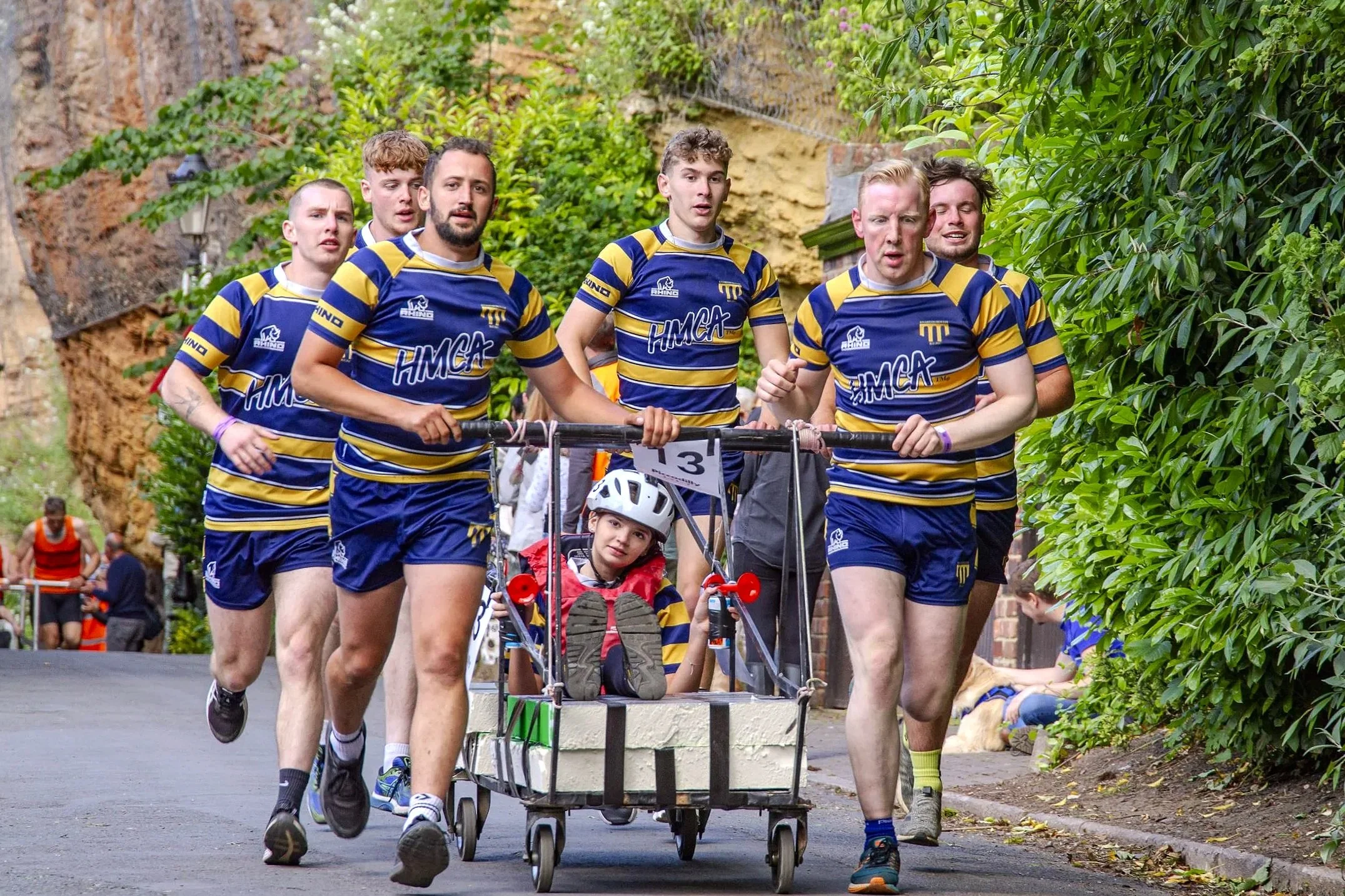 A group of six young men in blue and yellow striped rugby jerseys running outdoors, pushing a cart with a young girl wearing a white helmet sitting inside.