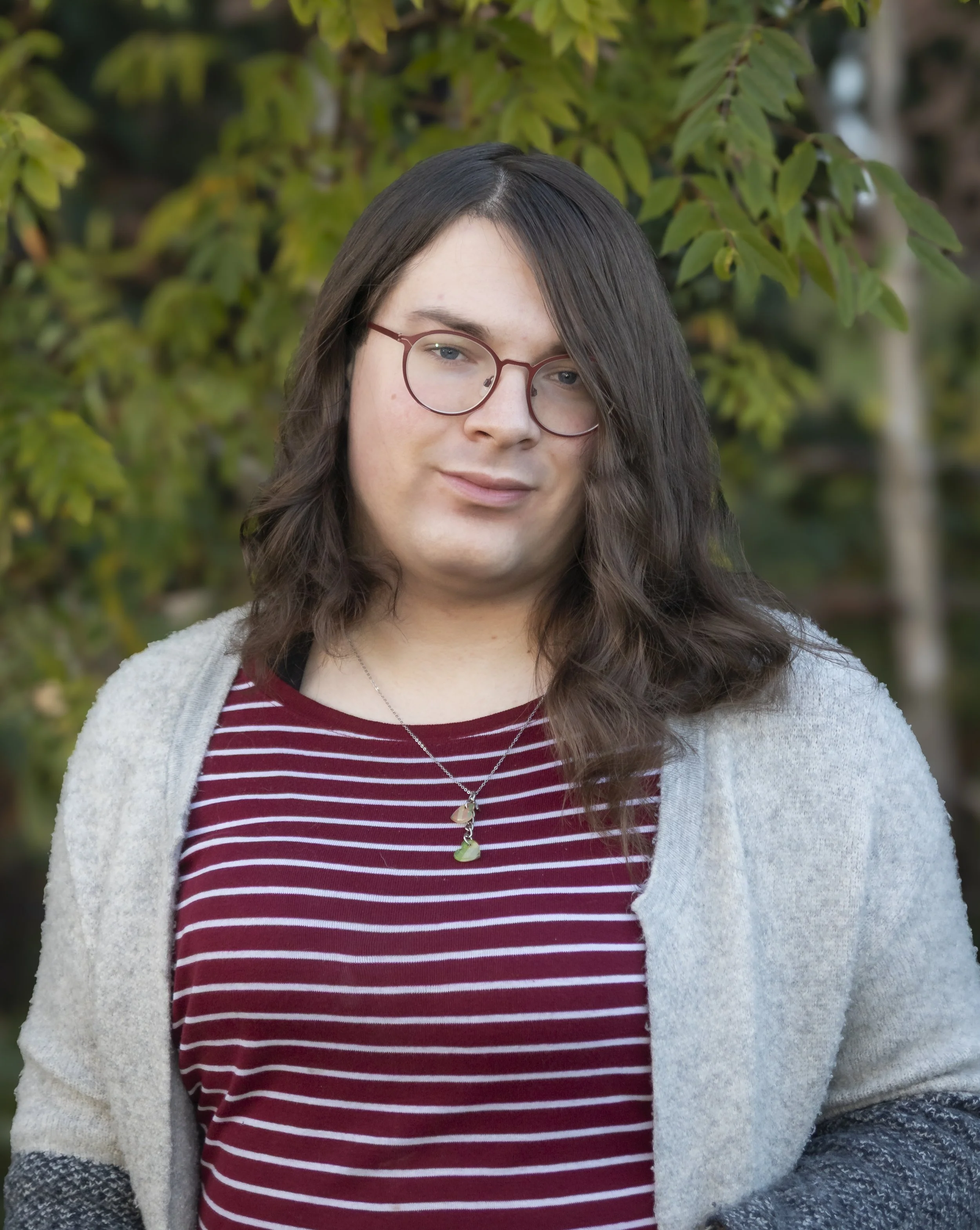 A young person with long dark hair, wearing glasses, a striped maroon shirt, a gray cardigan, and a necklace, standing outdoors in front of green foliage.