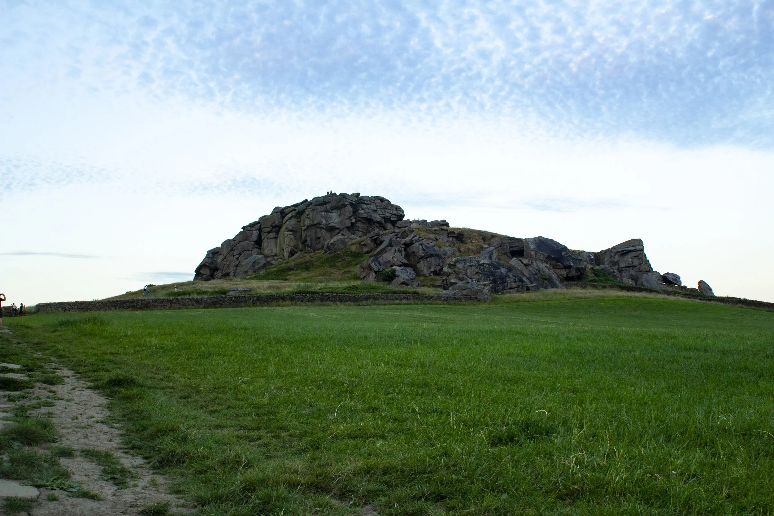 A grassy landscape with a large rocky hill or formation under a partly cloudy sky.