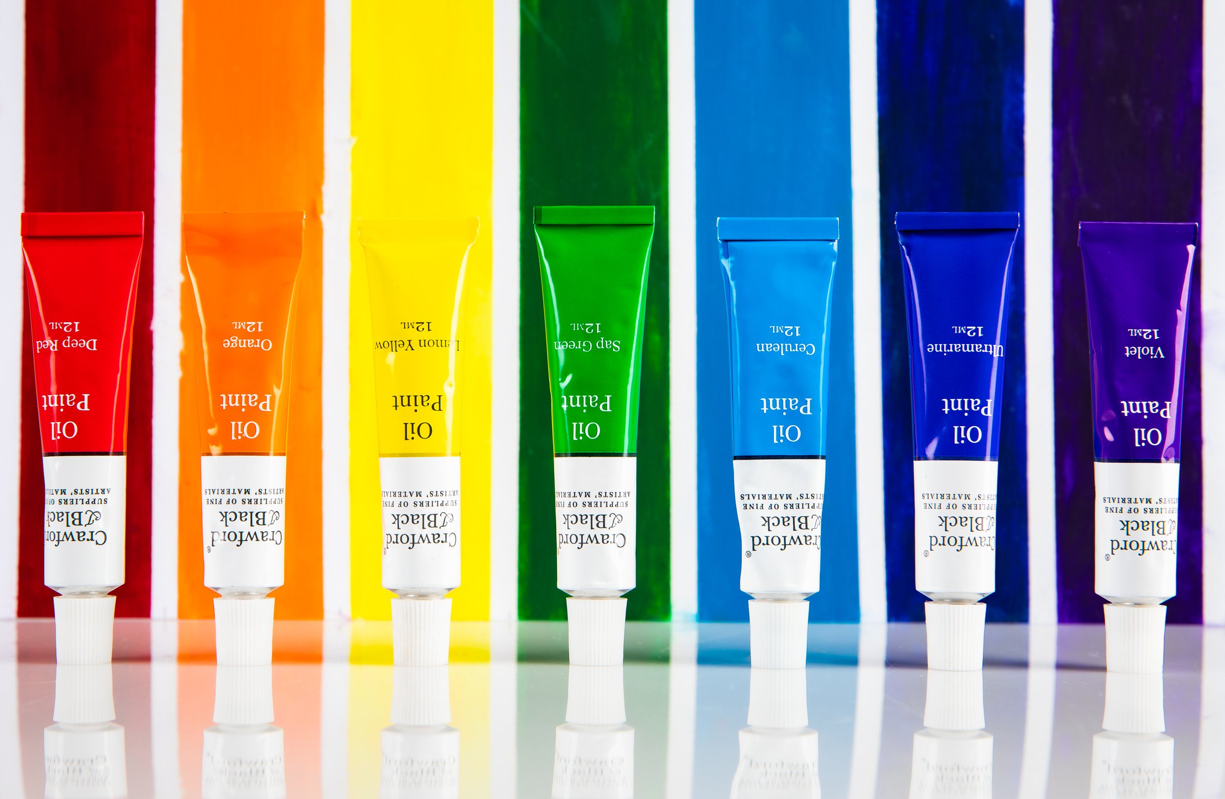 A row of seven tubes of watercolor paint in red, orange, yellow, green, blue, dark blue, and purple, with their corresponding color backgrounds reflected on a shiny surface.