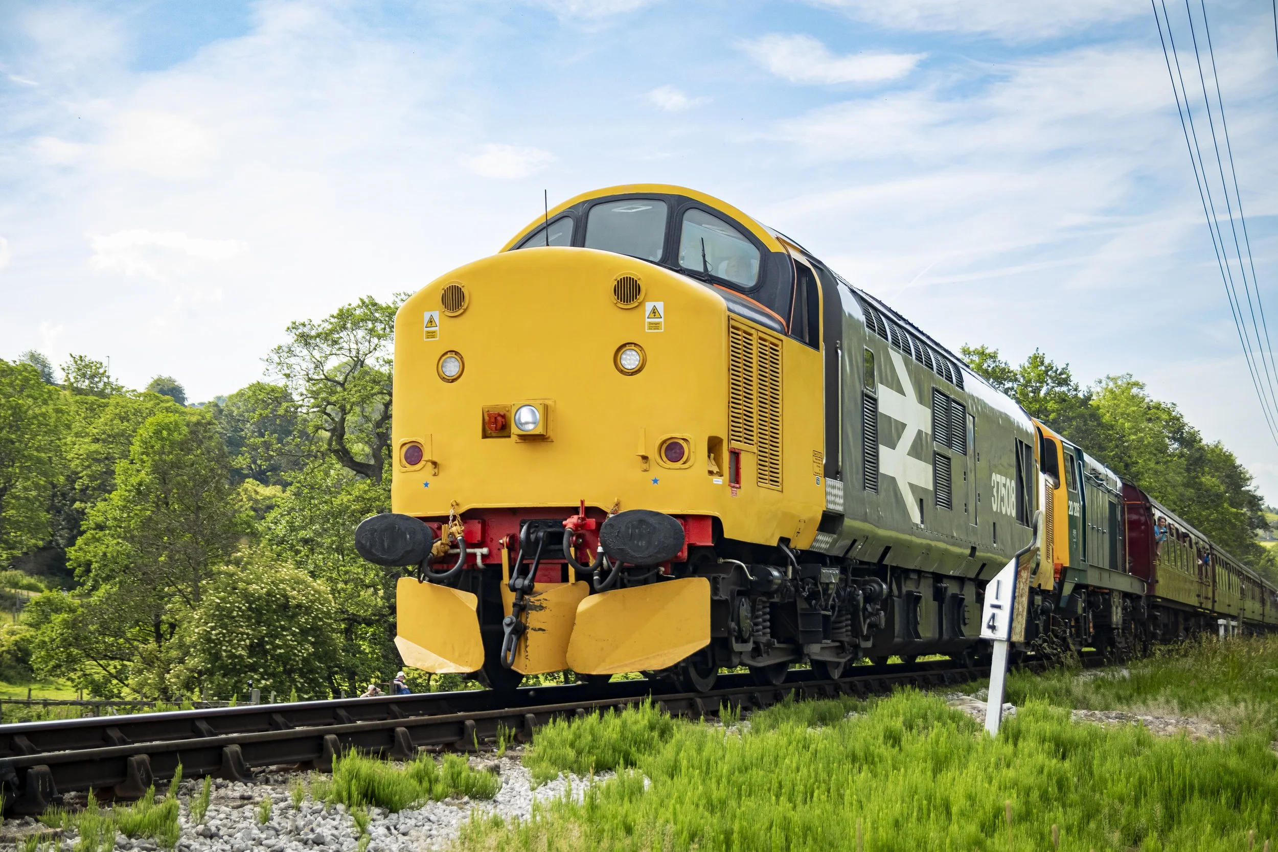 A vintage yellow and green train traveling on a railway track through a green countryside with trees and a blue sky.