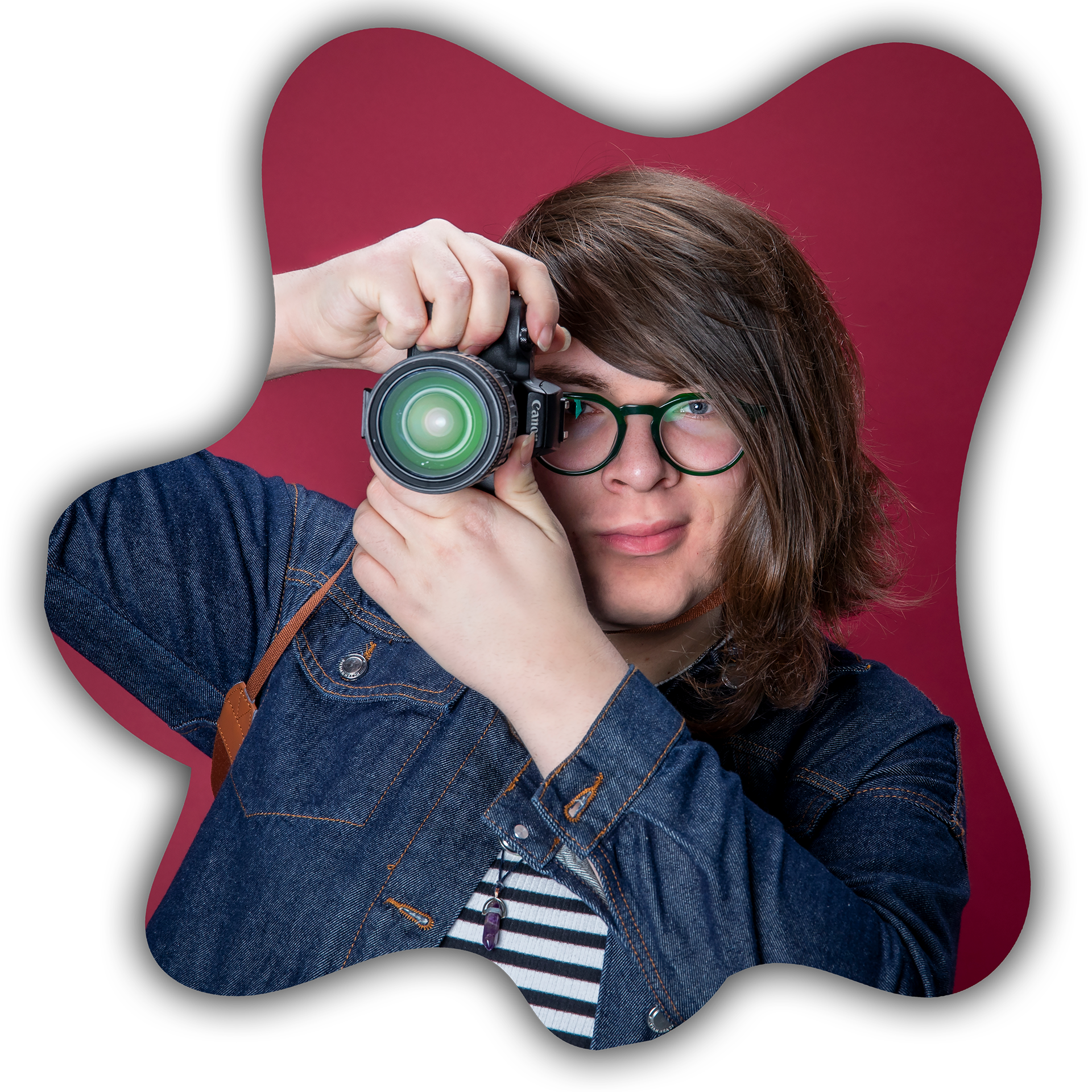 Person with shoulder-length hair and green glasses holding a camera to their eye, wearing a denim jacket and a striped shirt, with a solid pink background.