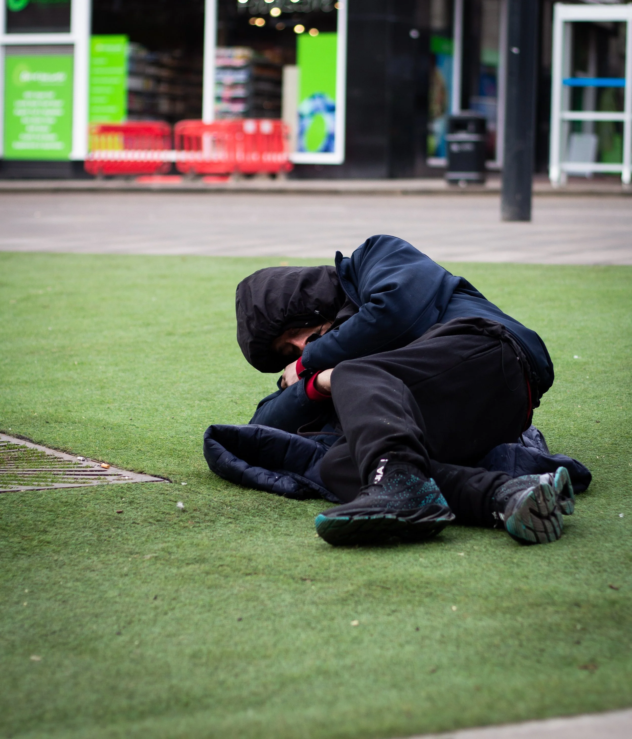 A person in a dark hooded jacket and black pants is sitting on the ground next to a patch of artificial green grass, hugging a person in dark clothing, on what appears to be a sidewalk or street outside a store.