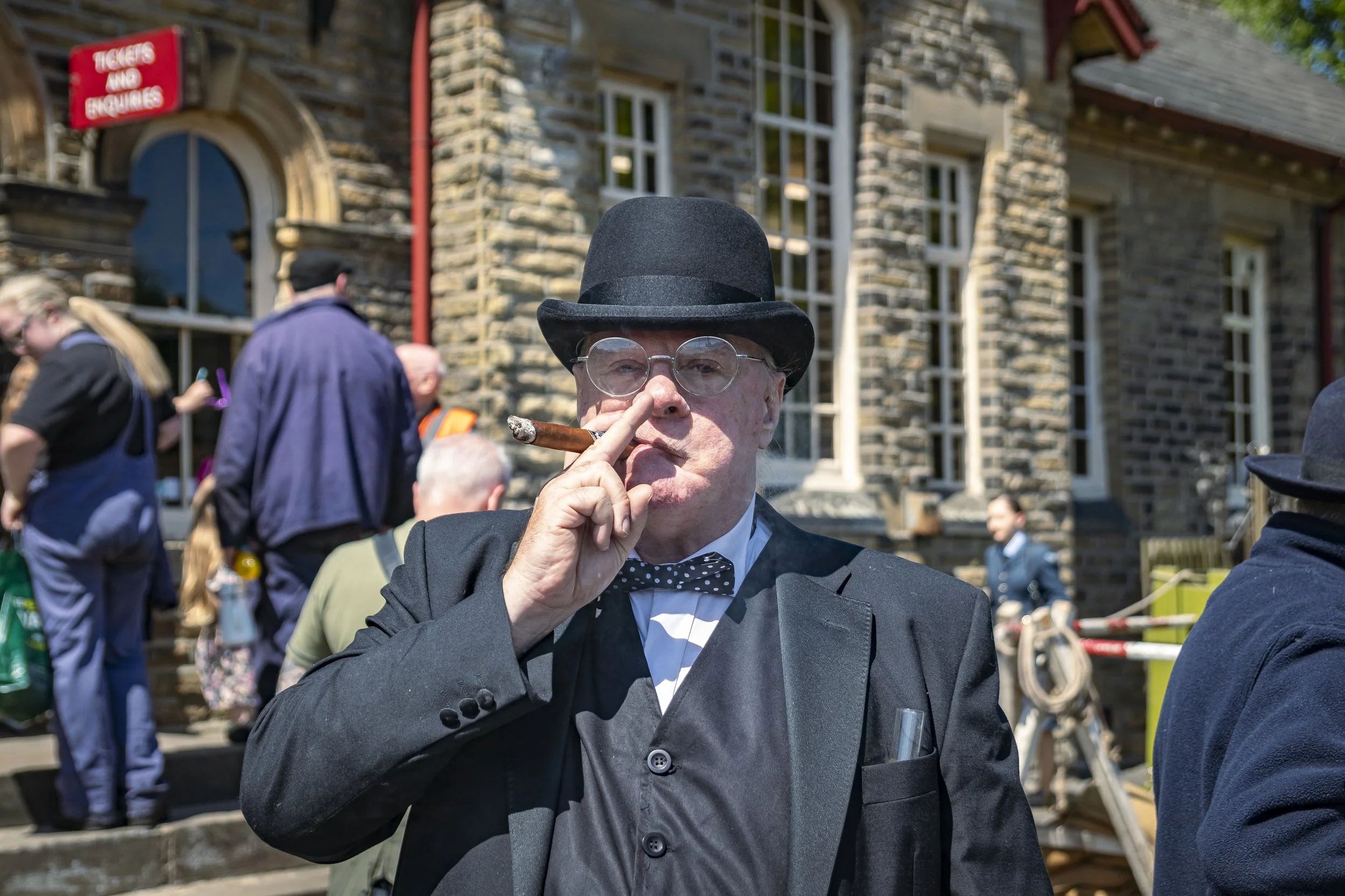 A man in a black suit, bow tie, and black hat holding a cigar to his mouth, standing outside a stone building with a 'Tickets No Enquiries' sign, in a crowd of people.