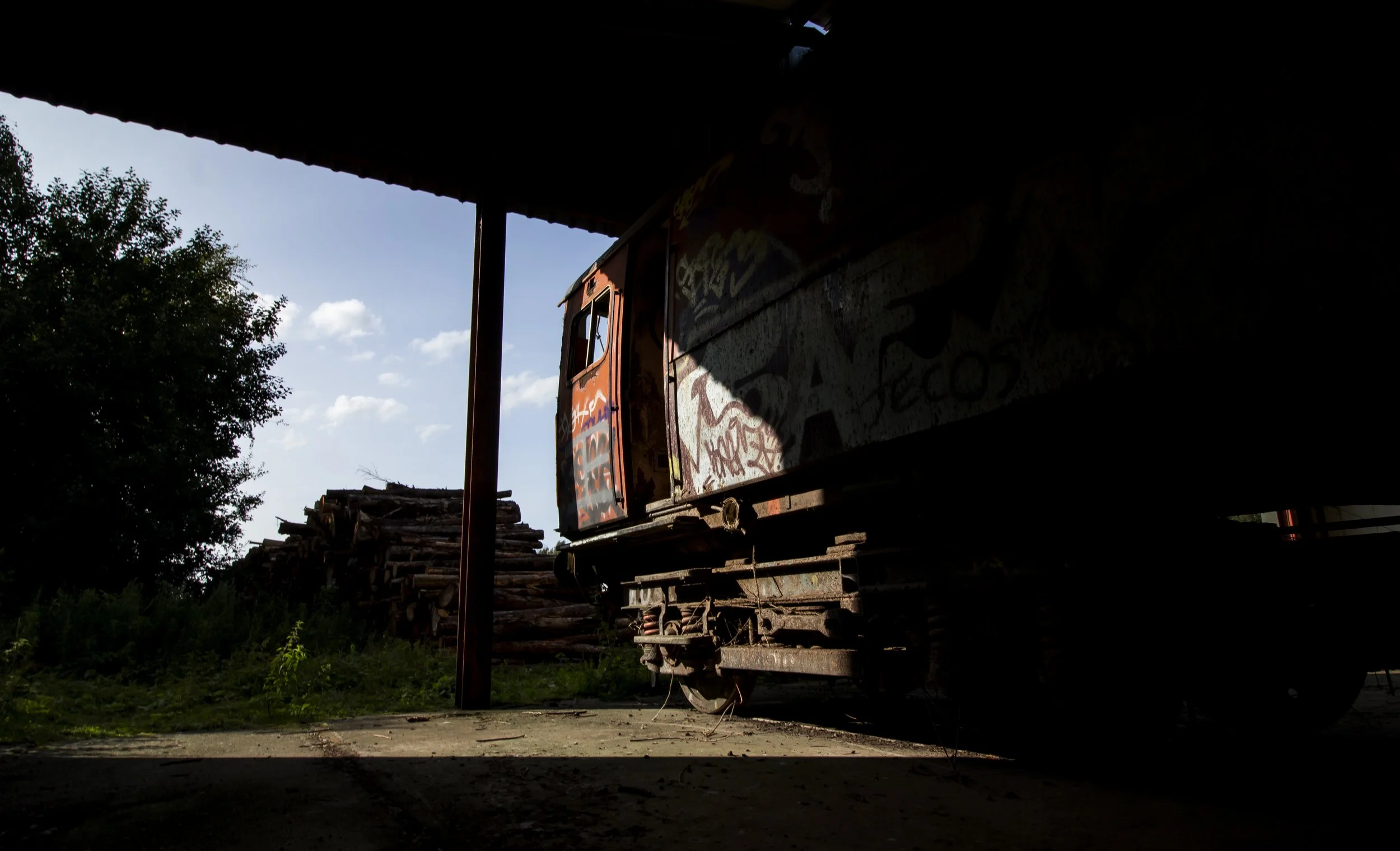 An old, graffiti-covered train car on a rusty track in a partially shaded outdoor setting, with a pile of stacked logs nearby and a tree against a partly cloudy sky.