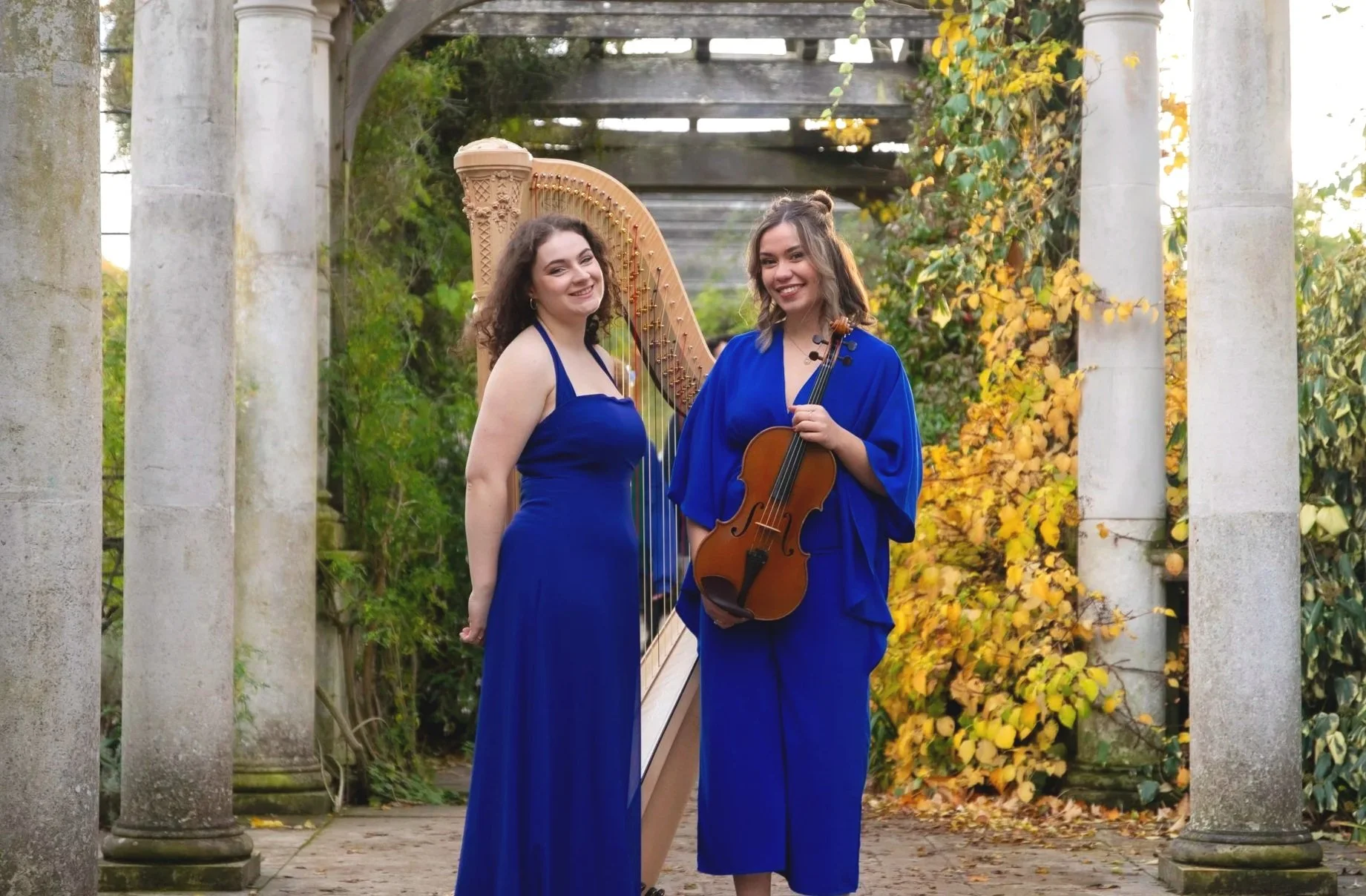 Two women in blue dresses holding musical instruments, a harp and a violin, standing outdoors among stone columns and autumn foliage.