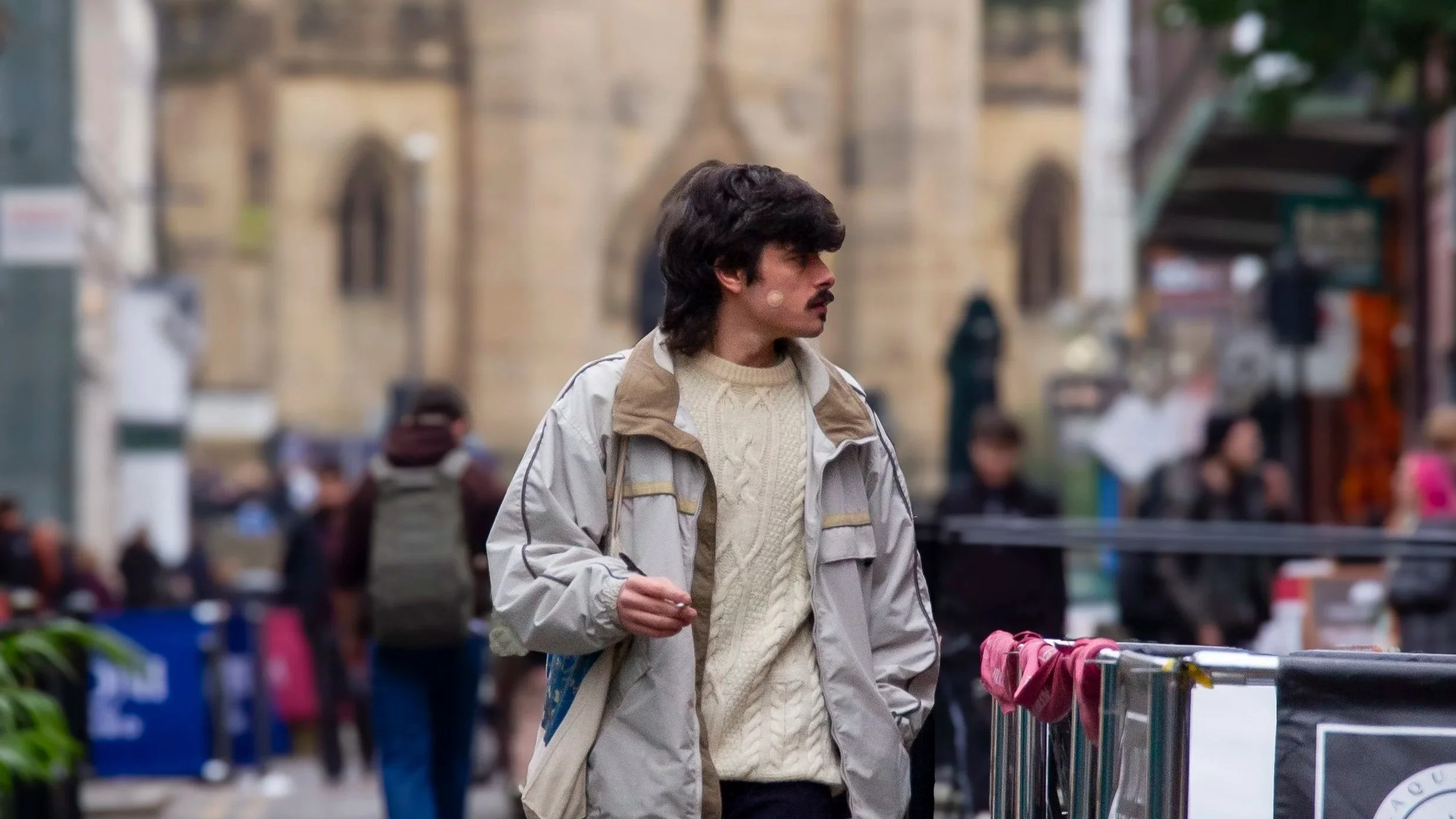 A man with dark, wavy hair and a mustache walking along a busy street with people in the background, wearing a beige and gray jacket over a cream-colored sweater.