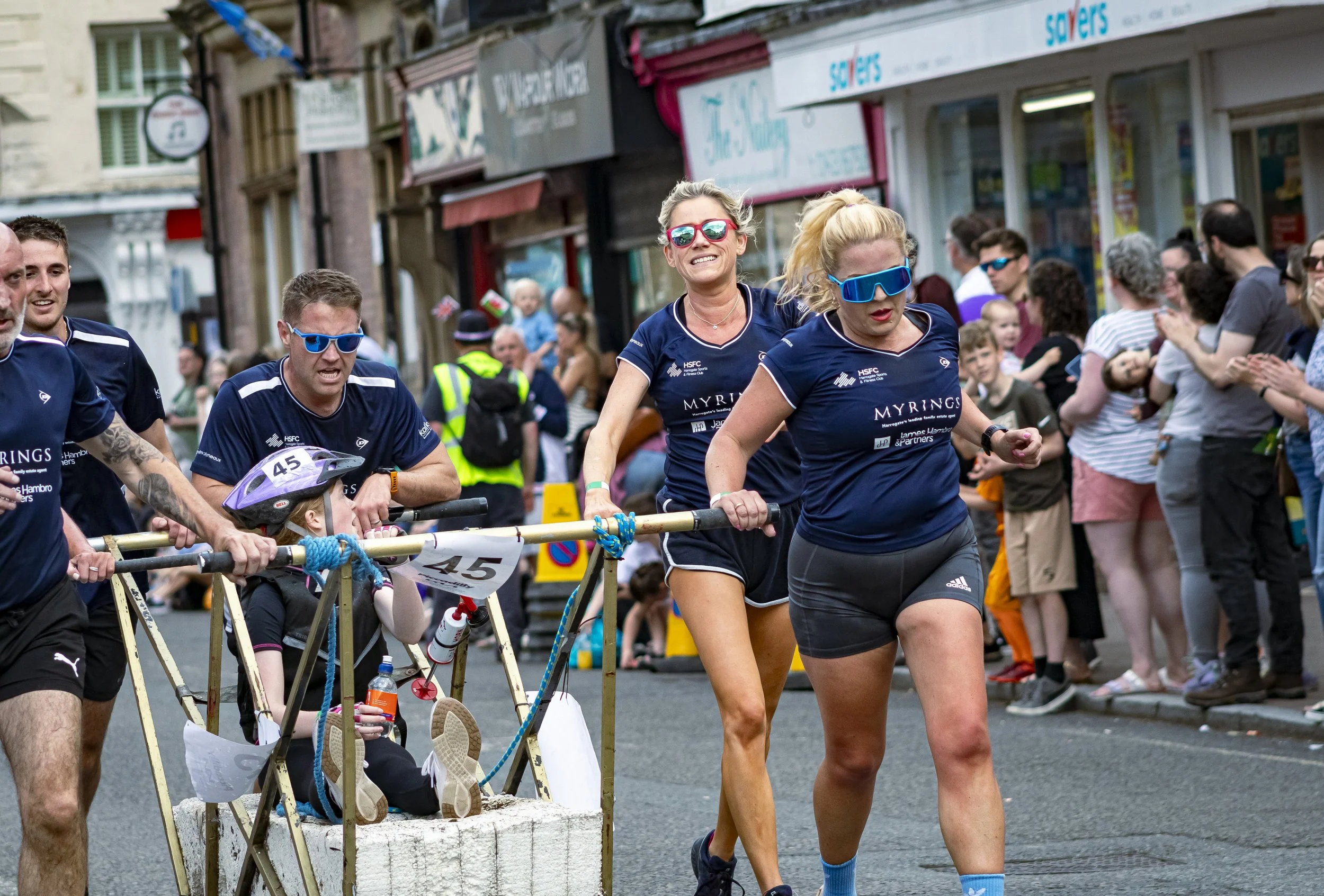 Participants in a marathon race, pushing a small contraption with a young person in a helmet, on a city street lined with spectators.