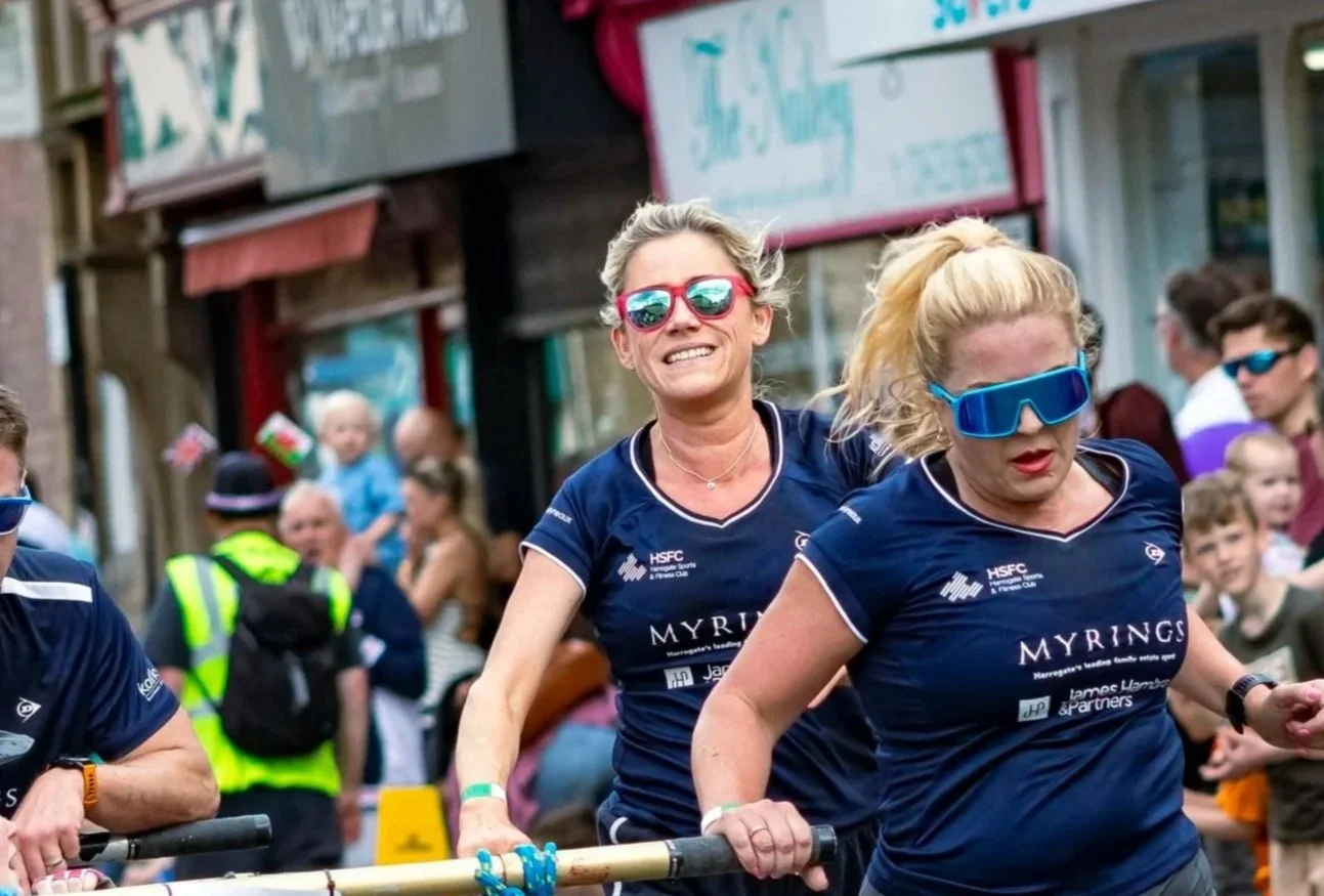 Two women wearing blue sports jerseys and futuristic sunglasses participating in a parade or event, with a crowd and storefronts in the background.