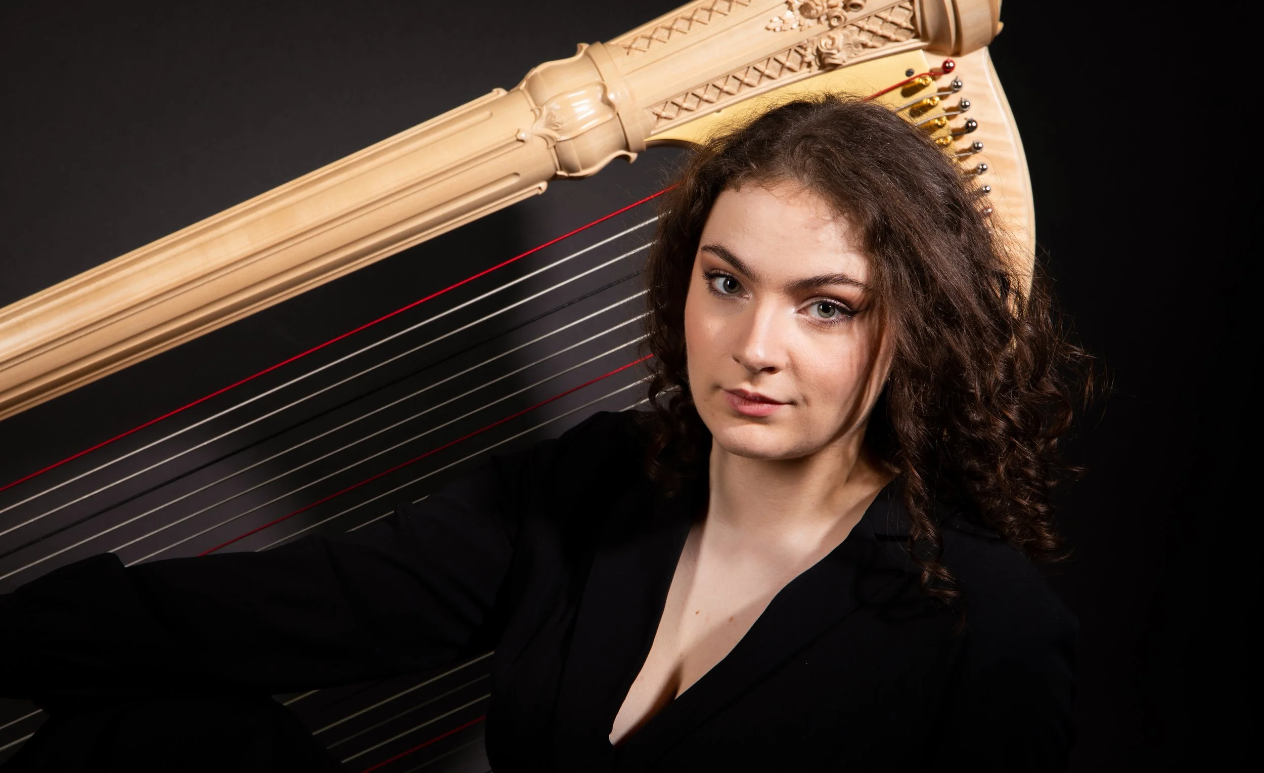 Young woman with curly hair sitting in front of a large harp, wearing a black top, against a dark background.