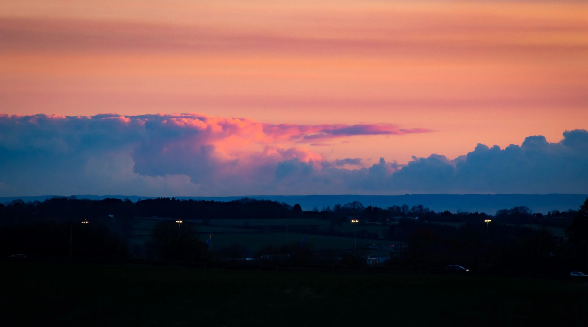 A sunset sky with pink and purple clouds over a dark landscape with trees, hills, and streetlights.