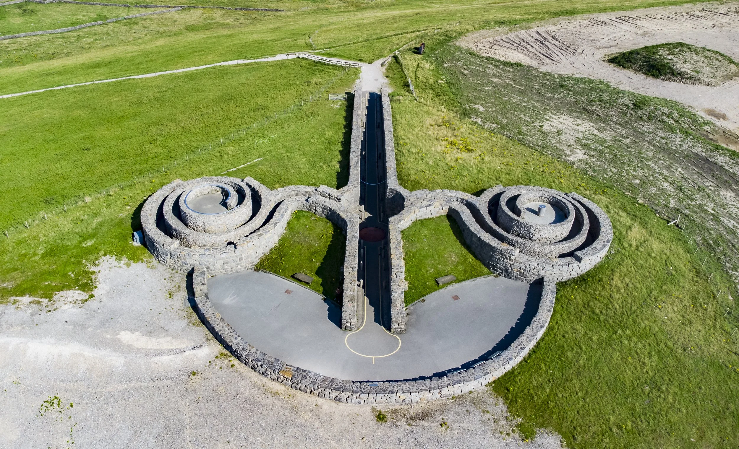 Aerial view of a large stone sculpture in a grassy area, consisting of two concentric spirals connected by a central divider, with a paved circle in front.
