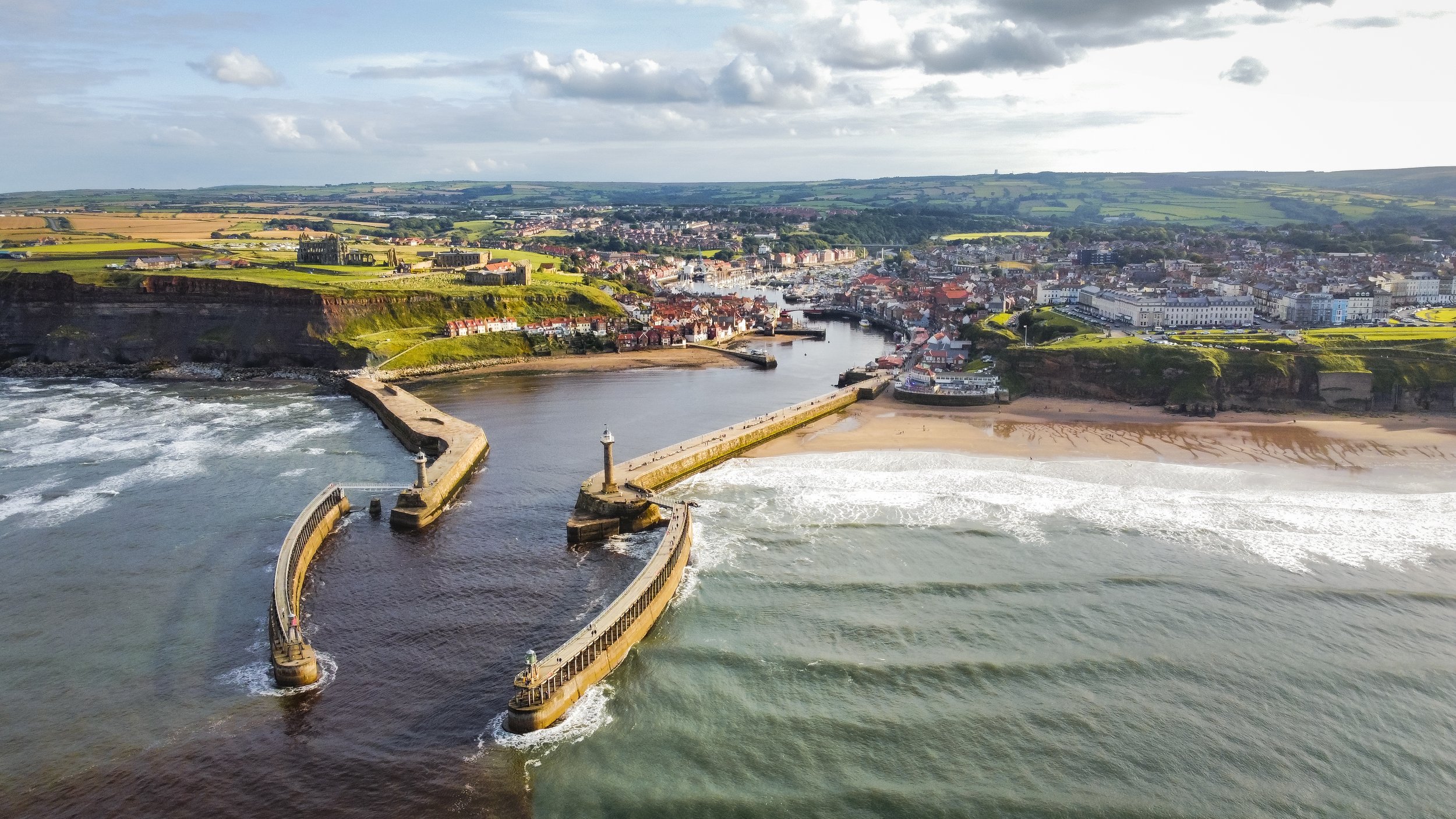 Aerial view of a coastal city with a harbor, a breakwater with a lighthouse, sandy beach, and surrounding green hills and buildings under a partly cloudy sky.