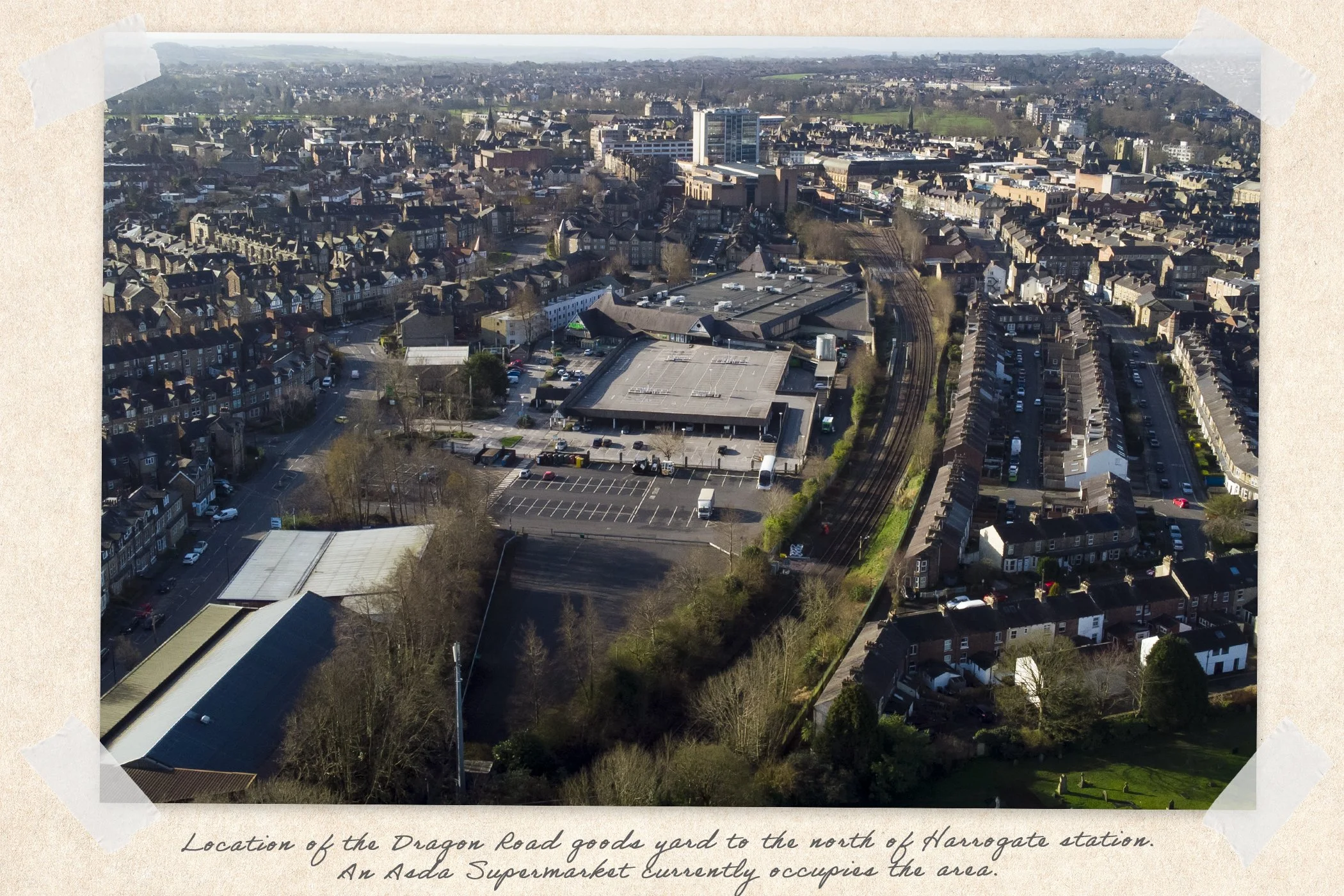 An aerial view of a town showing a supermarket with a large parking lot, surrounded by residential houses, and train tracks running nearby. The area is busy with streets and vehicles. The scene is in daylight with clear weather.