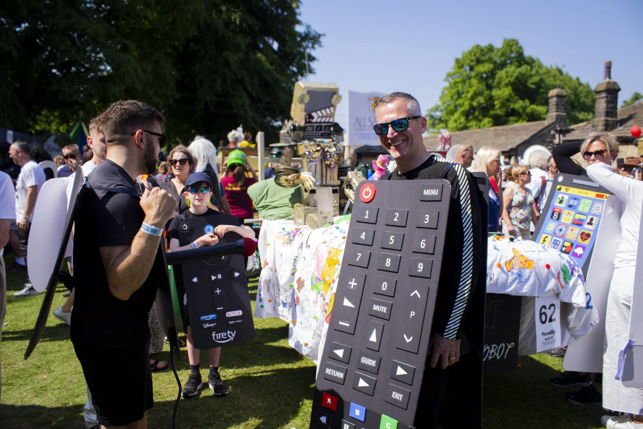 People attending an outdoor event with some wearing costumes and accessories. A man is smiling and holding a large remote control costume. Other people are browsing tables with items for sale, and the scene is sunny with green trees in the background.