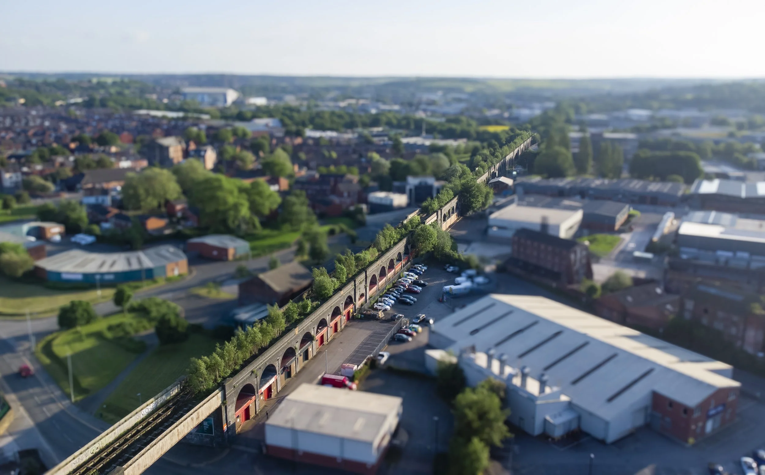 Aerial view of a city showing a viaduct with trees, buildings, and parking lots, with a blurred cityscape in the background under a partly cloudy sky.