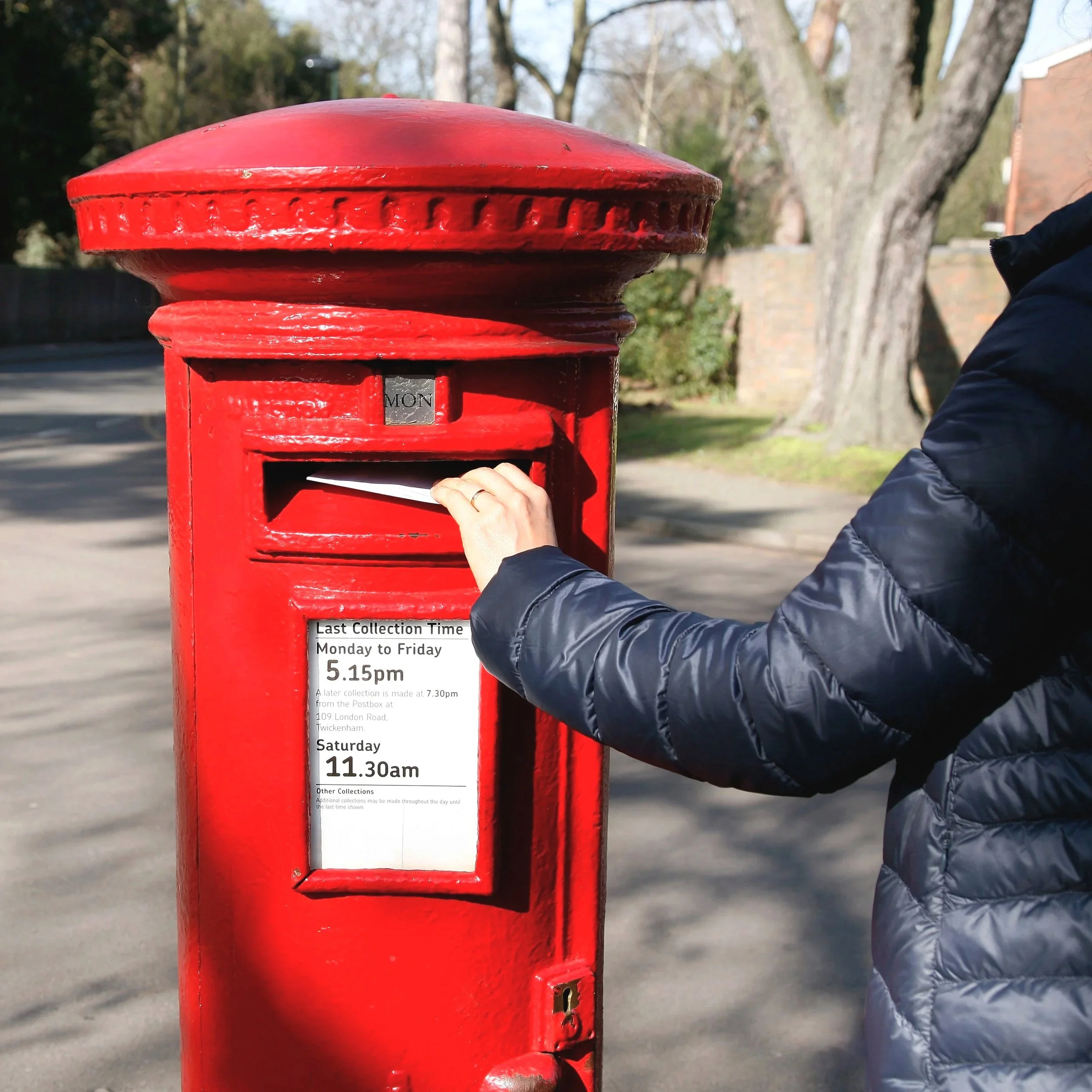 Person in a navy blue jacket delivering mail into a red mailbox on a sunny street with trees and brick wall in the background.