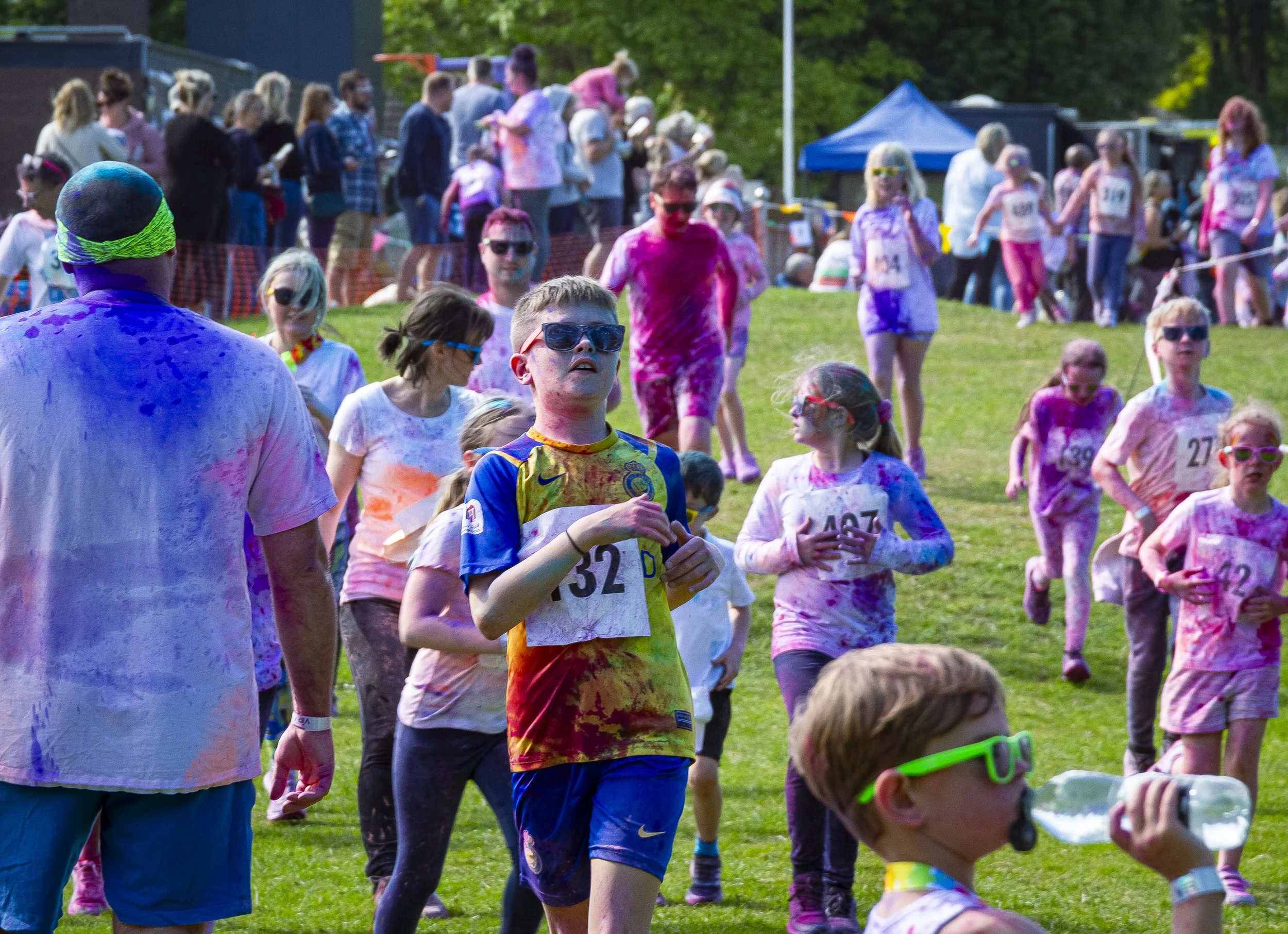 Children and adults participating in a color run, with colorful powder on their clothes and faces, outdoors on a grassy field with tents and spectators in the background.