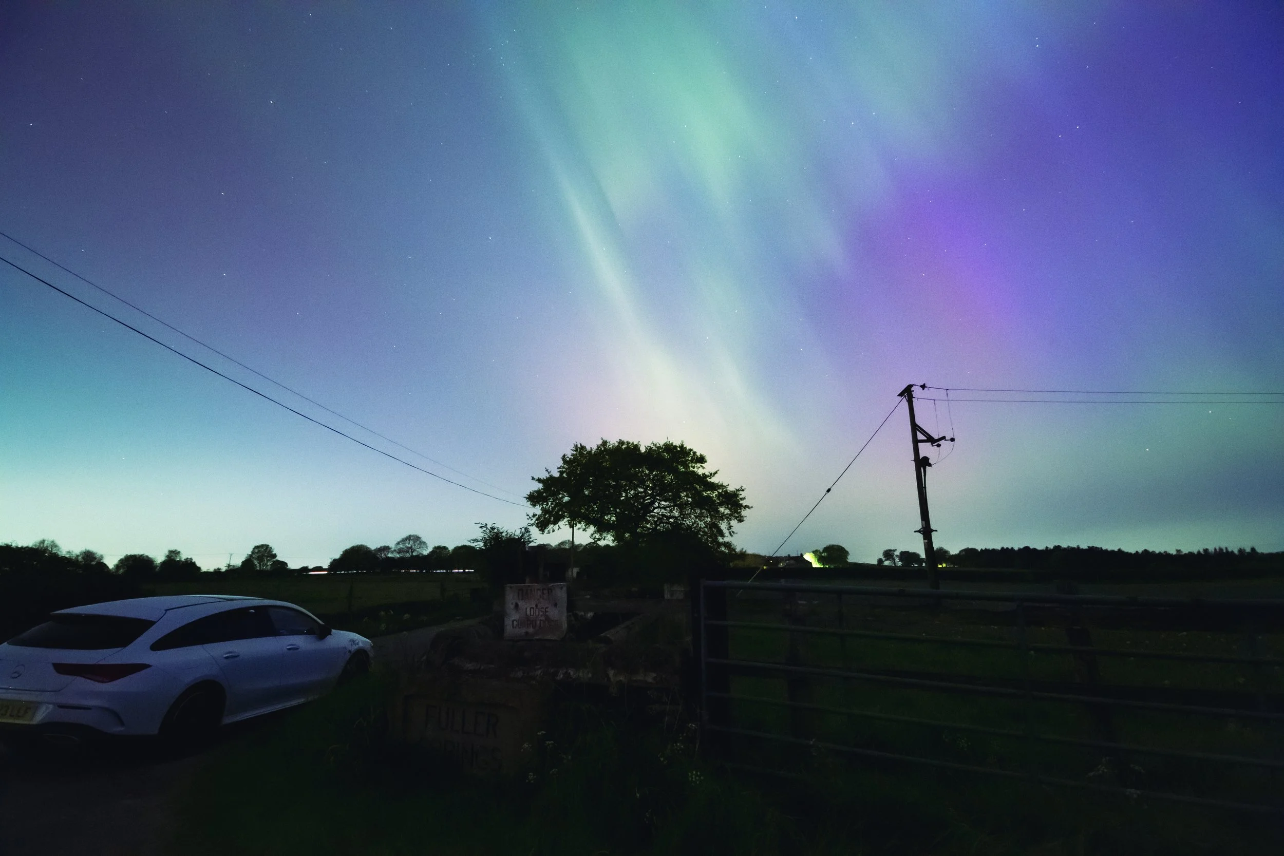 Night sky with colorful aurora borealis, a white car parked on the grass, and power lines and trees in the background.