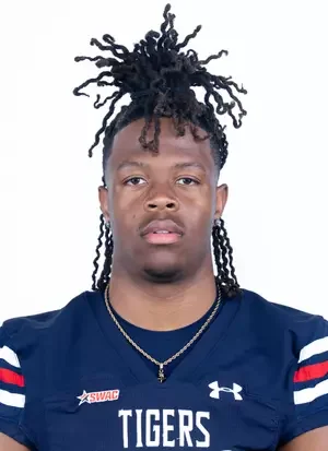 Portrait of a young man with styled dreadlocks wearing a navy sports jersey with red, white, and blue stripes and a gold chain.