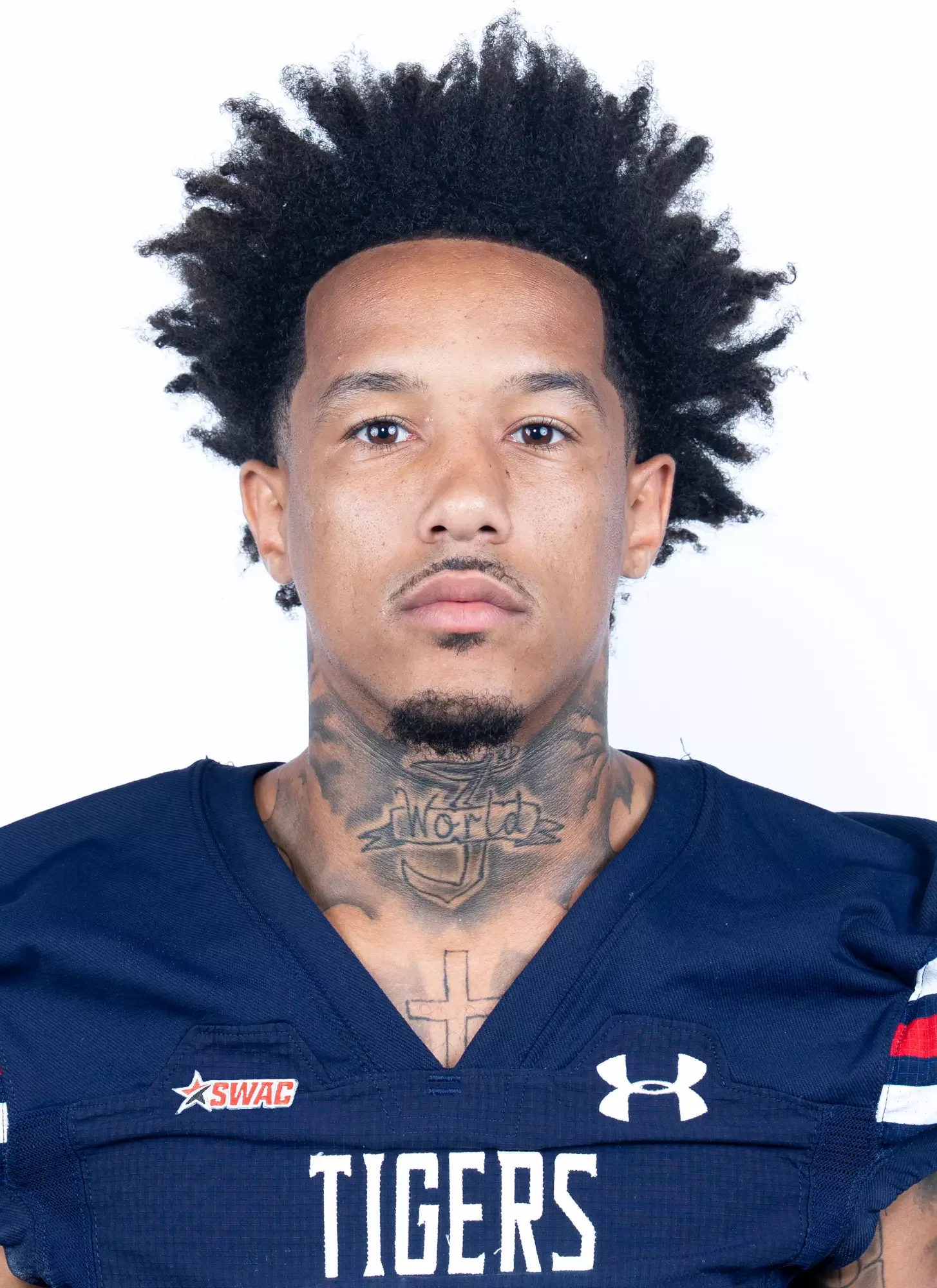 Close-up portrait of a young African American male athlete with tattoos on his neck, wearing a navy blue jersey with 'TIGERS' written on it, and the Under Armour logo. He has curly hair and a serious expression.