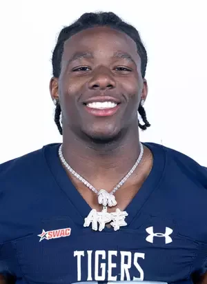 Smiling young man with braided hair wearing a navy sports jersey with 'TIGERS' and Under Armour logos, a diamond necklace, and a WWE Championship belt around his neck against a white background.