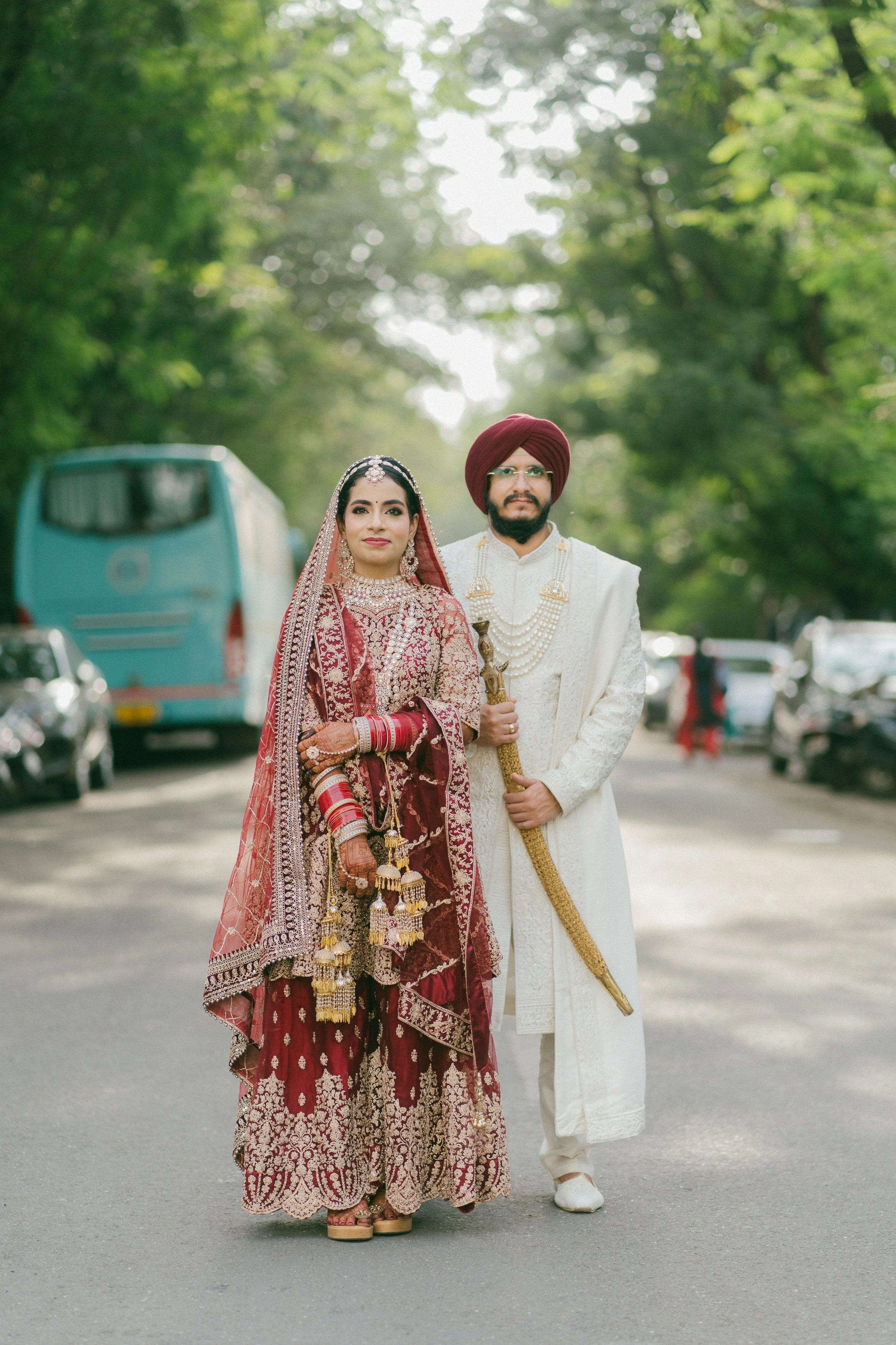 A couple dressed in traditional Indian wedding attire standing on a street lined with cars and buses, with greenery in the background.