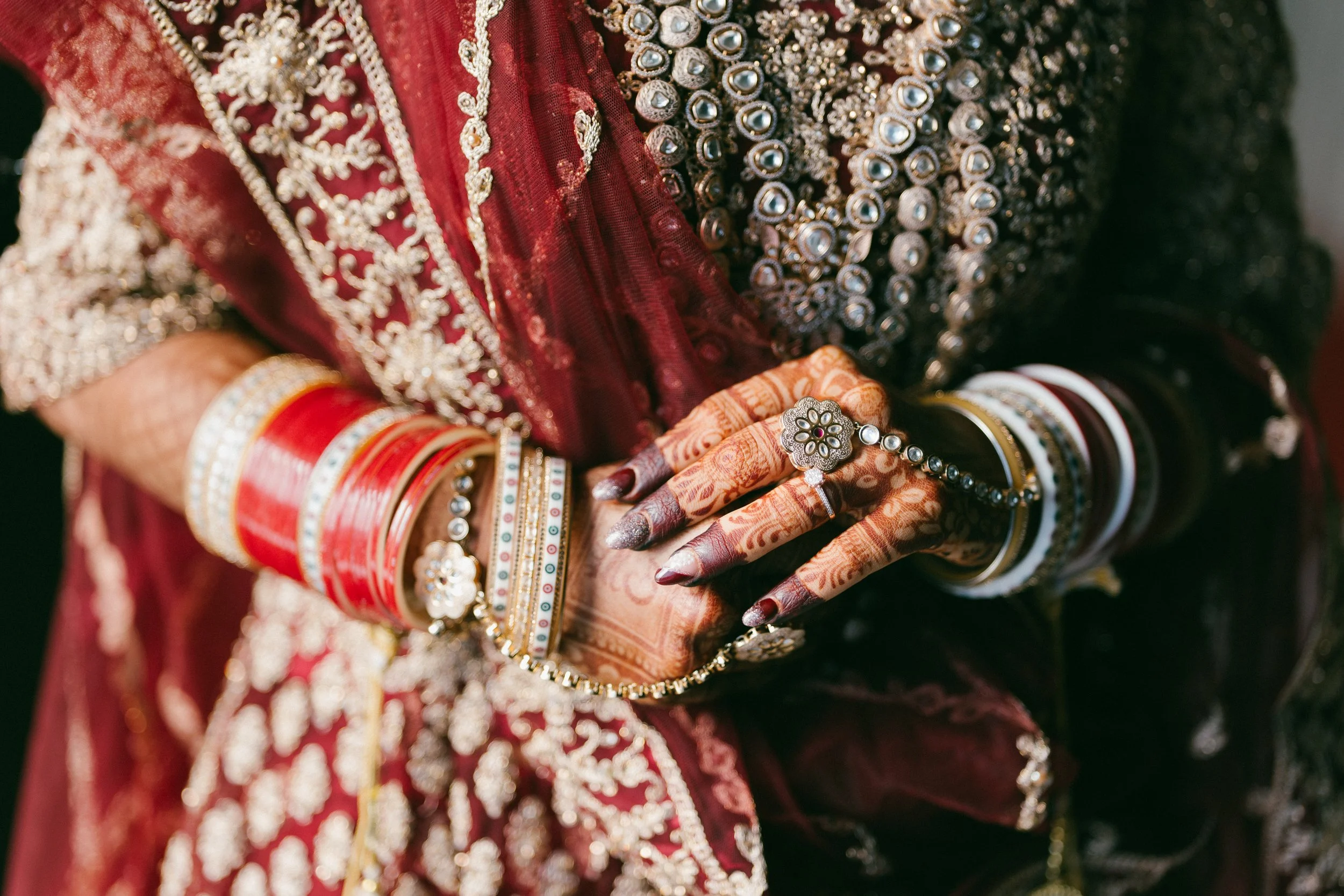 Close-up of a woman wearing traditional Indian bridal attire, featuring ornate bangles, rings, and a heavily embroidered dress. Her hands are decorated with henna designs.