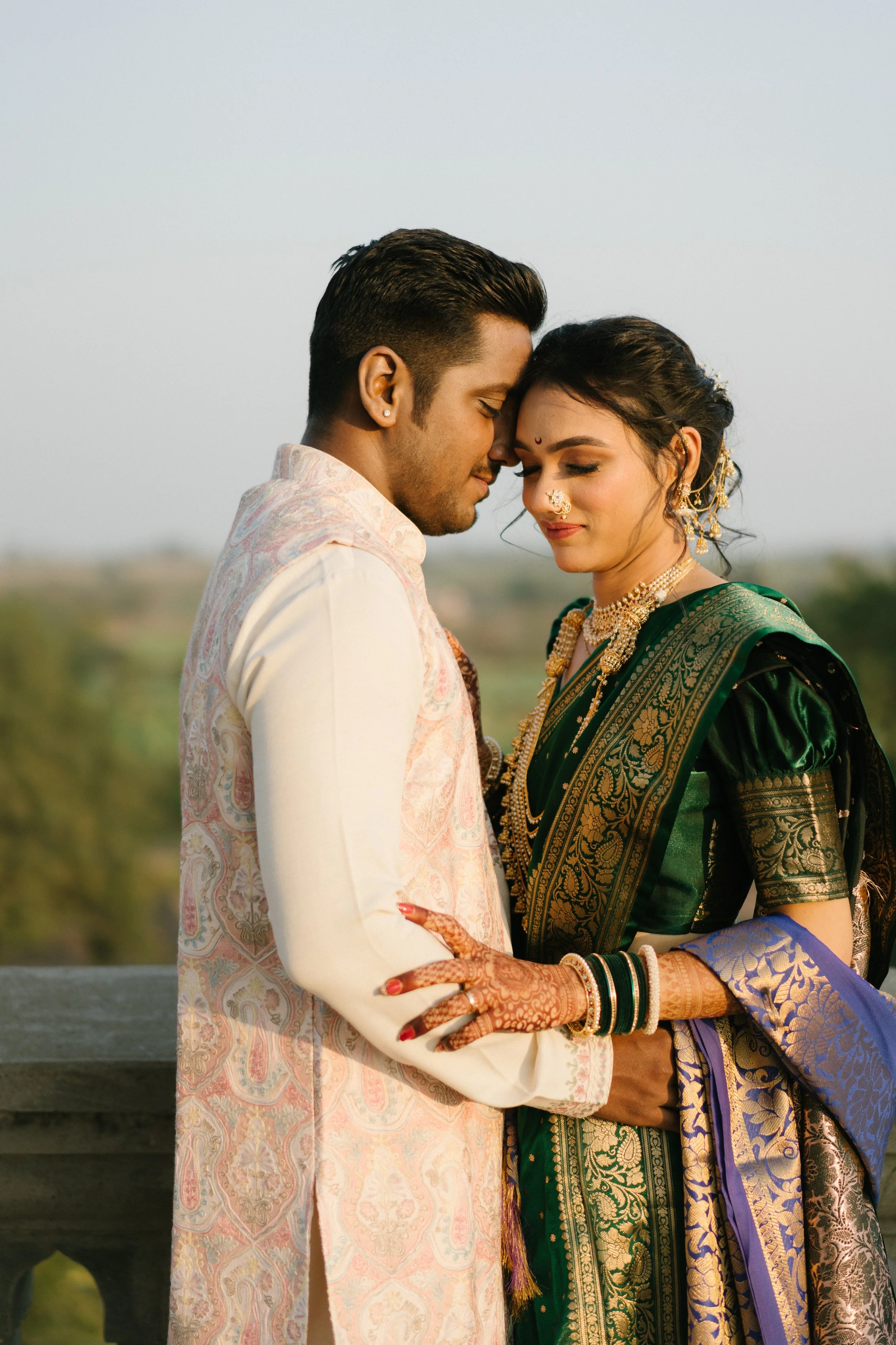 A couple dressed in traditional Indian wedding attire, standing close with foreheads touching, outdoors with a blurred landscape background.