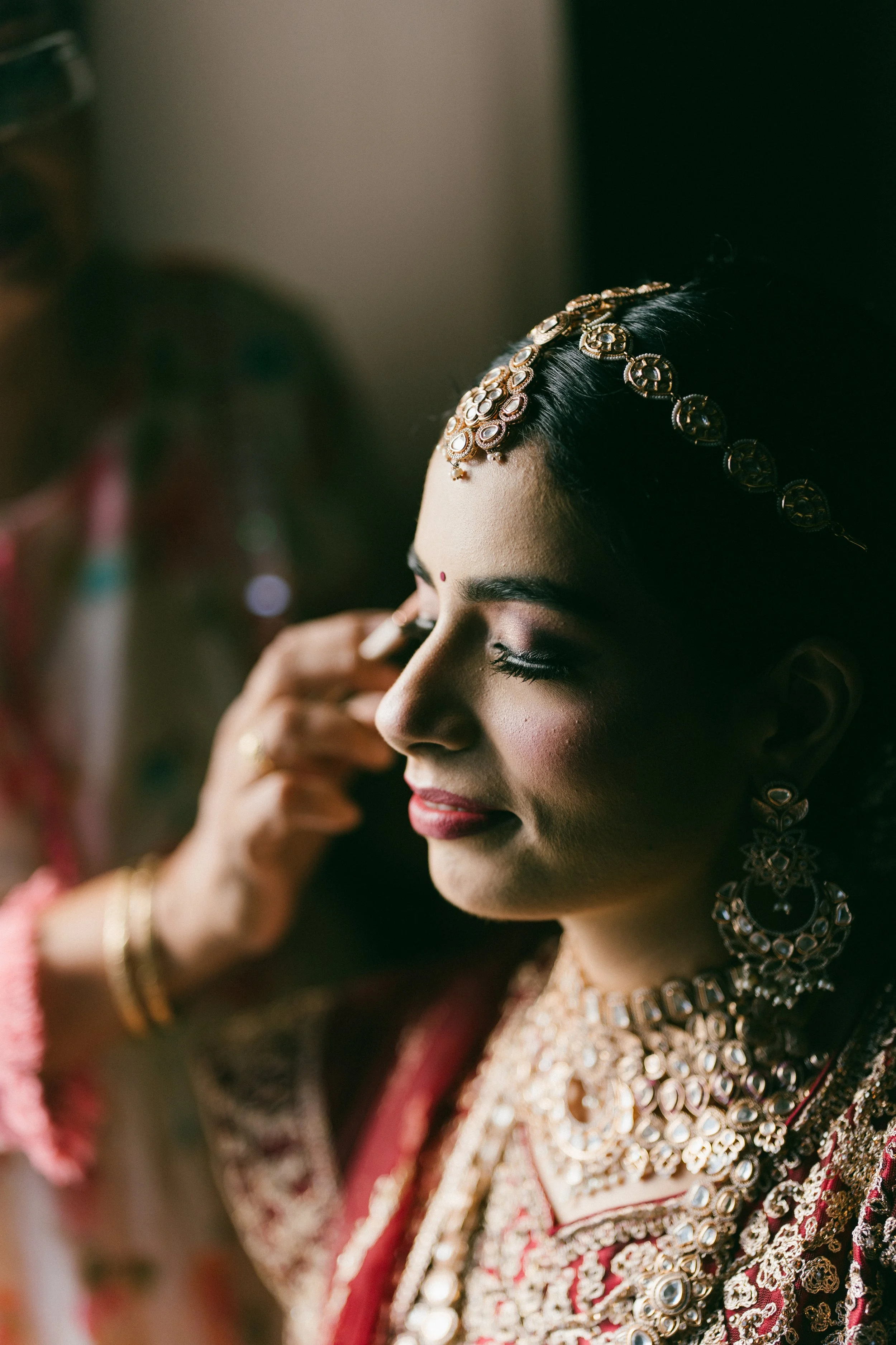 A woman with closed eyes, wearing traditional Indian jewelry and attire, having her makeup applied by a woman whose face is not visible.