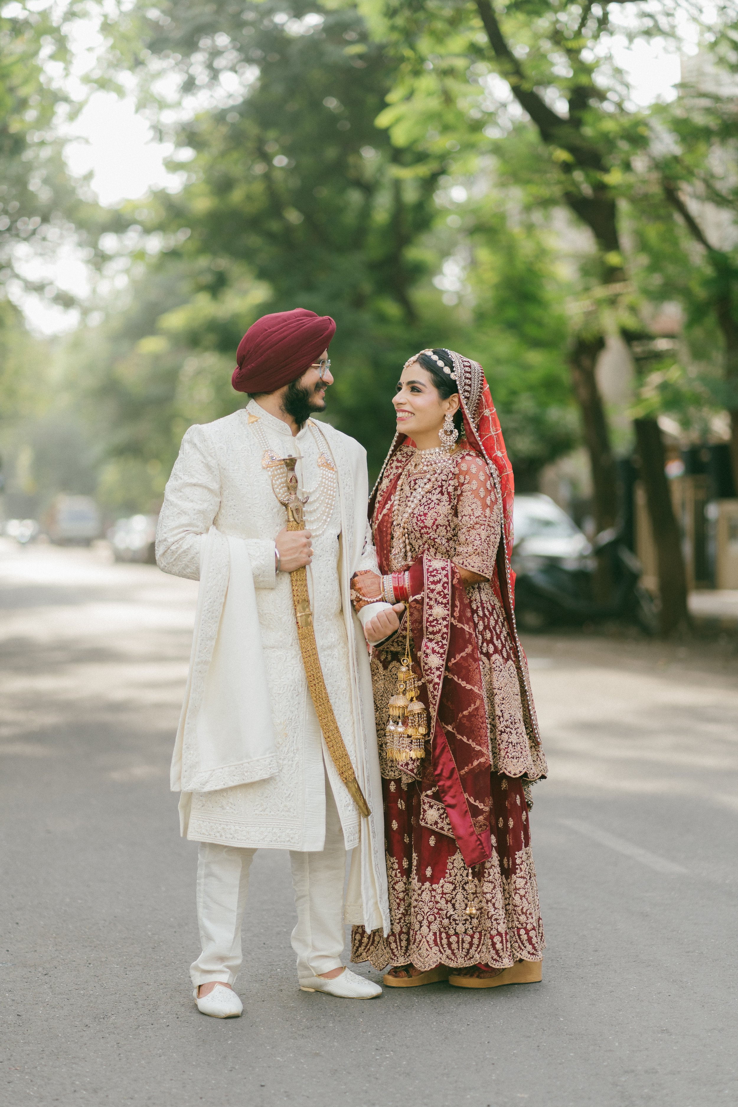 A newly married Indian couple in traditional wedding attire standing on a tree-lined street, smiling at each other.