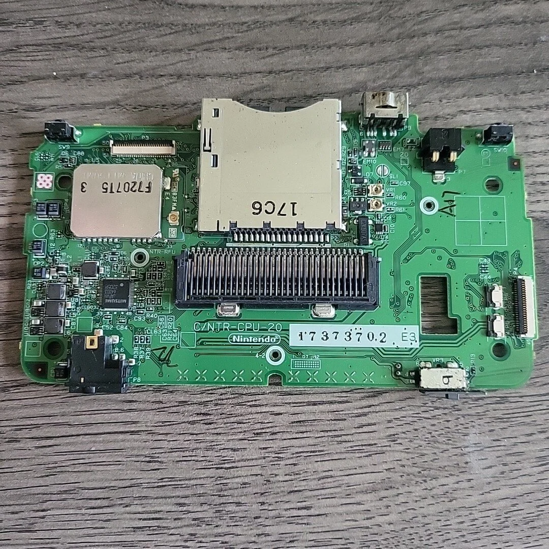 Green Nintendo Switch motherboard with various chips, connectors, and a metal shield, placed on a wooden surface.