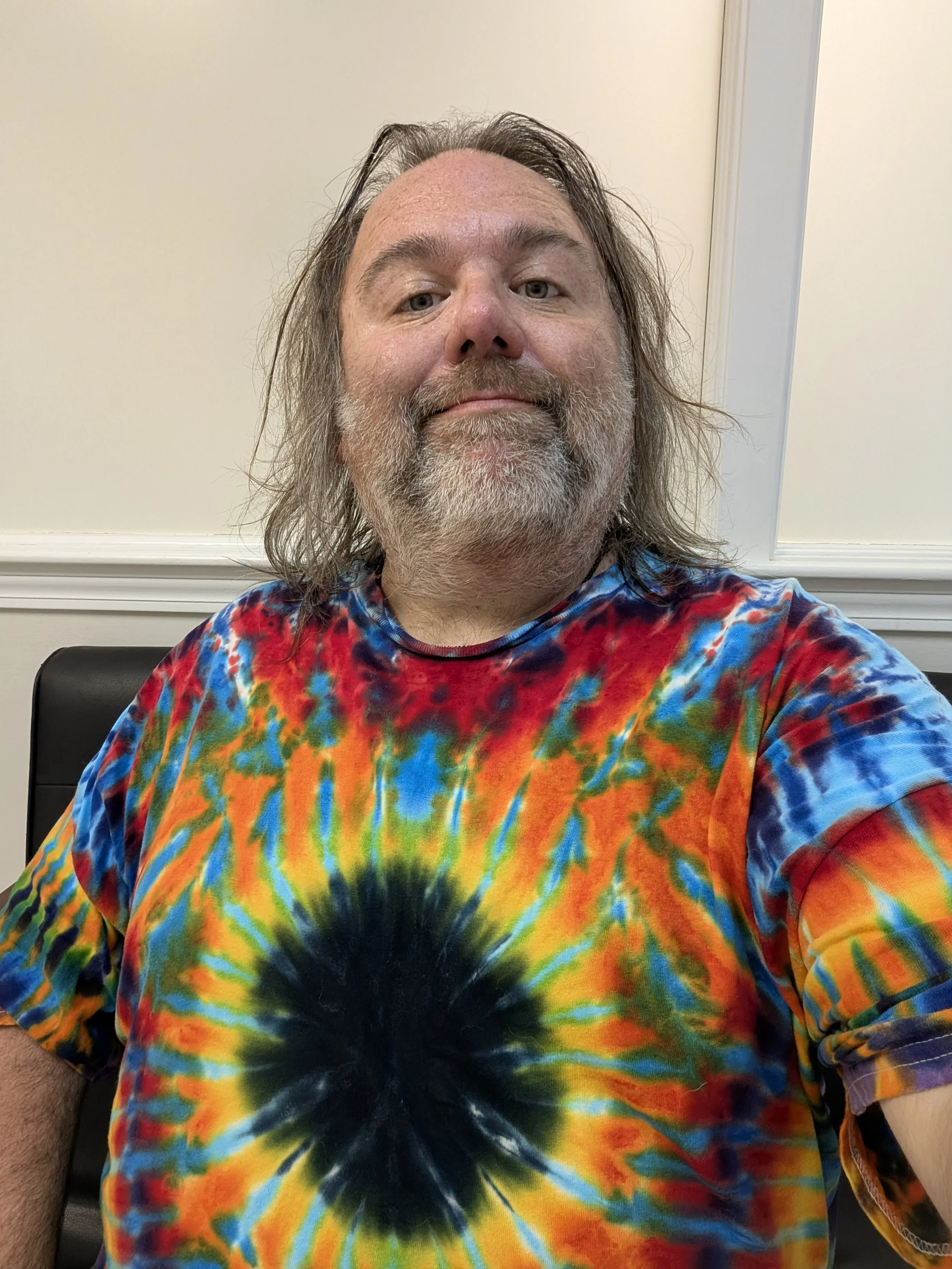 A man with shoulder-length gray hair and a beard takes a selfie, wearing a brightly colored tie-dye T-shirt with a black and yellow pattern at the center, in front of a plain cream-colored wall with white trim.