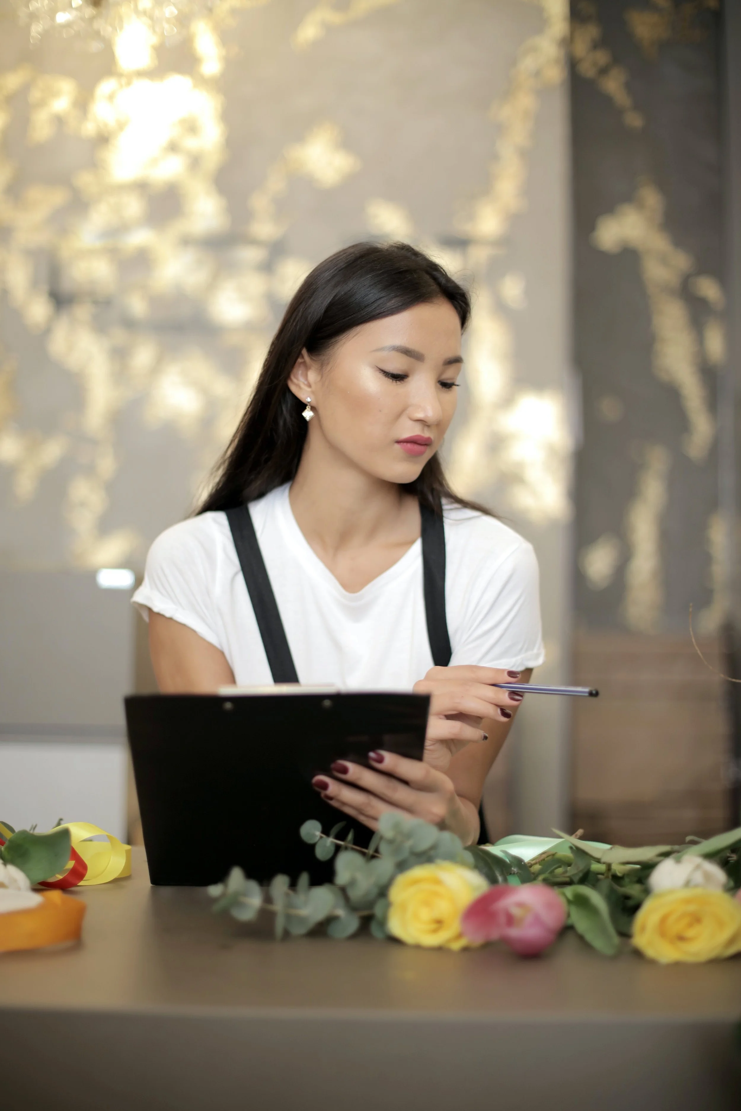 A woman with dark hair, wearing a white t-shirt and earrings, sitting at a table with flowers, holding a black notebook and a pen, in a room with a blurred background. She represents a person who wants help for anxiety in PA.